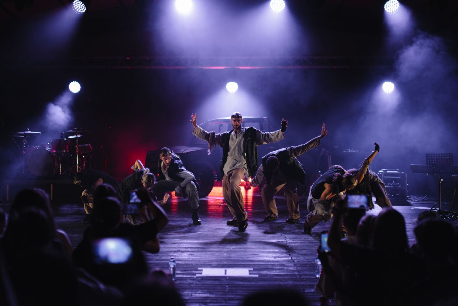 Performer with arms raised surrounded by dancers on a dramatic stage with blue and red lighting.