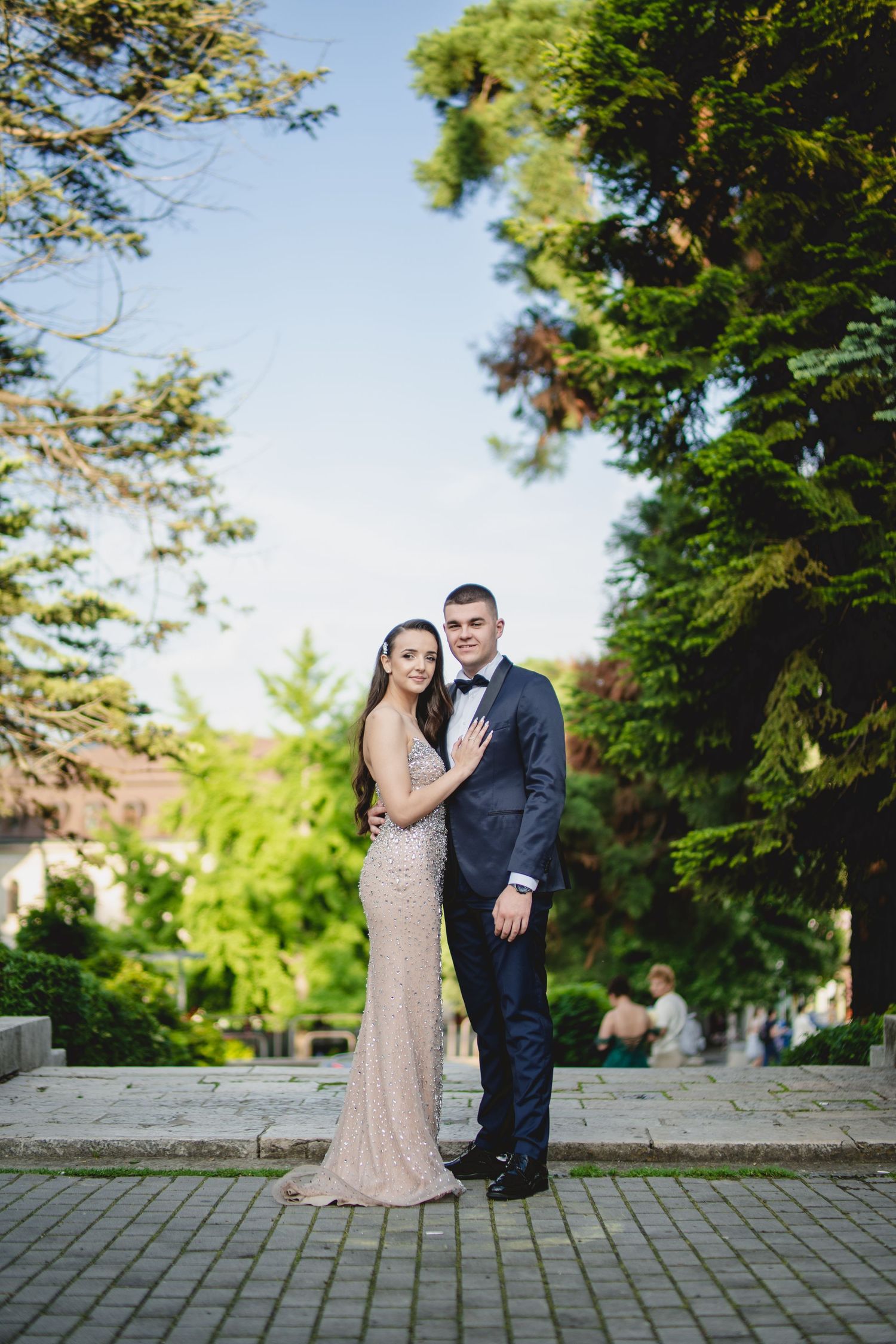 Couple in formal attire embracing outdoors, woman in sequin gown and man in navy suit.