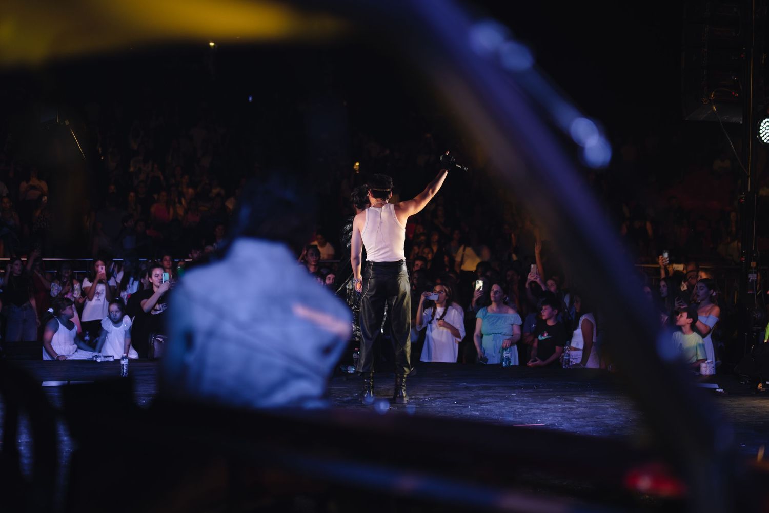 Performer on stage waving to a large crowd at an outdoor night concert viewed through a car window.