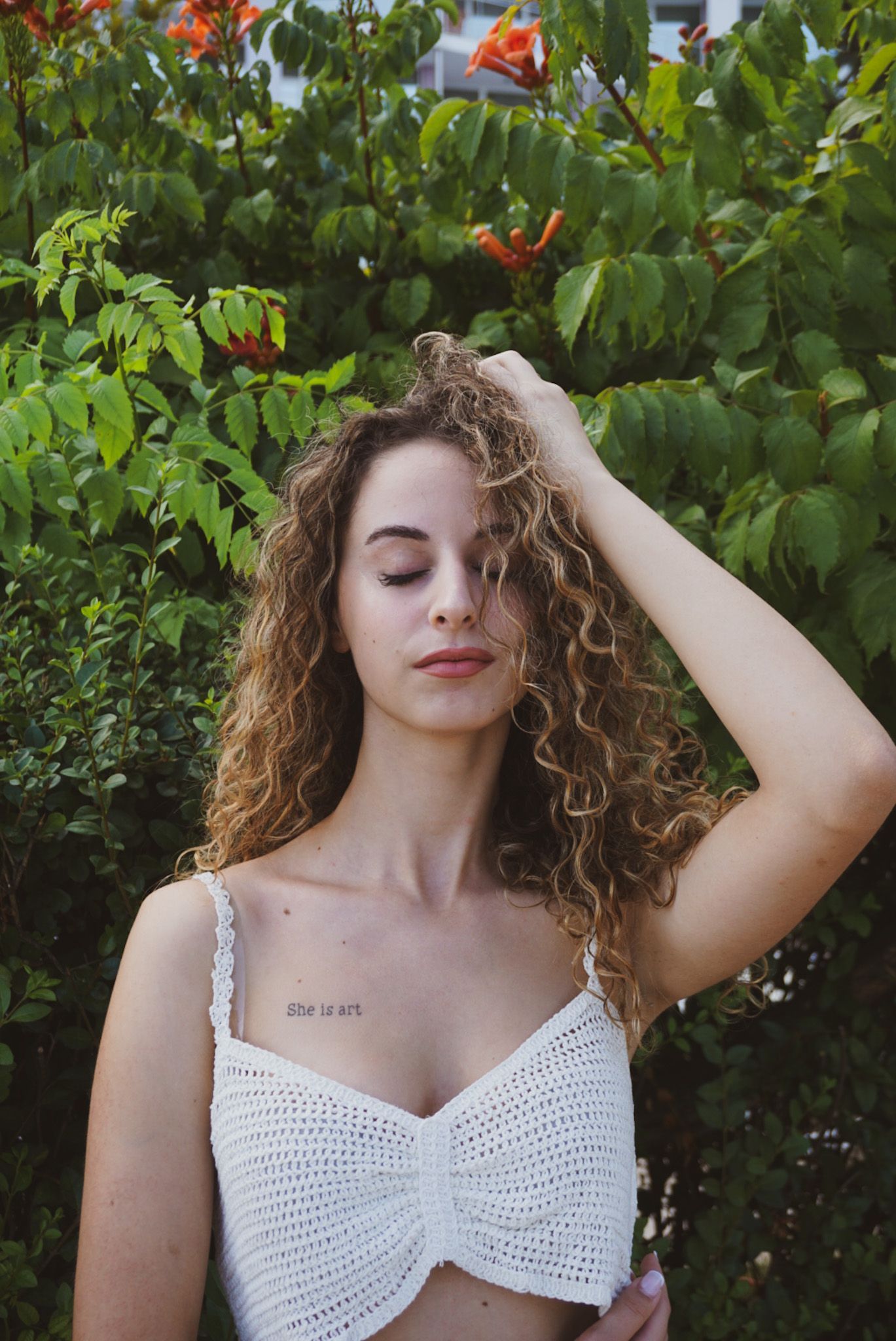 Young woman with curly hair and white crochet top standing near blooming orange flowers outdoors.