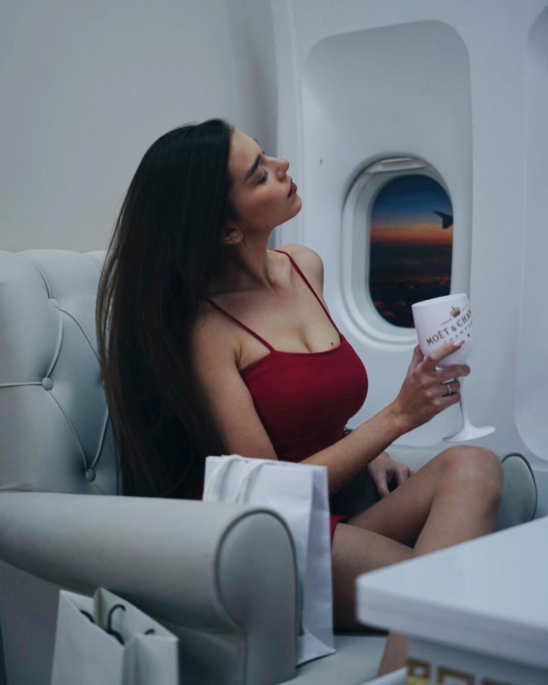 Elegant woman in red dress sipping champagne in luxury private jet seat by sunset window.