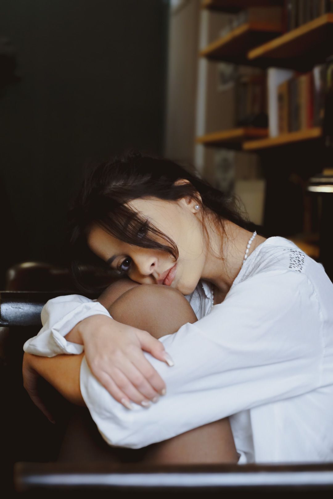 Brunette woman in white blouse resting head on arms at wooden table in cozy library setting.