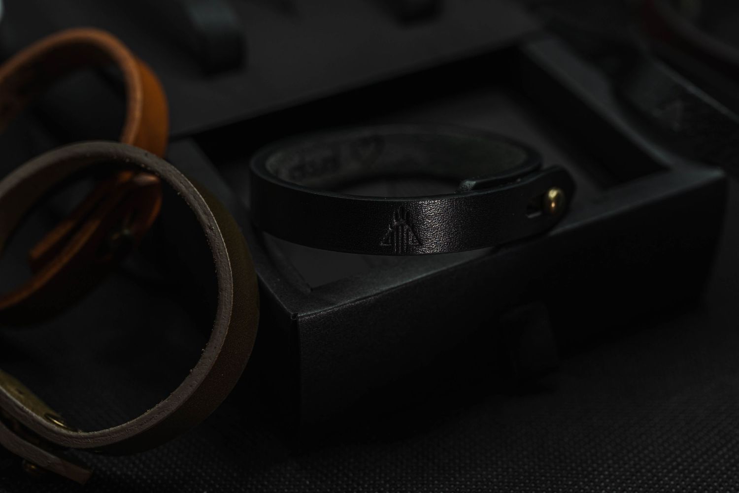 Black leather bracelet with embossed logo displayed on dark gift box surrounded by other leather bracelets.