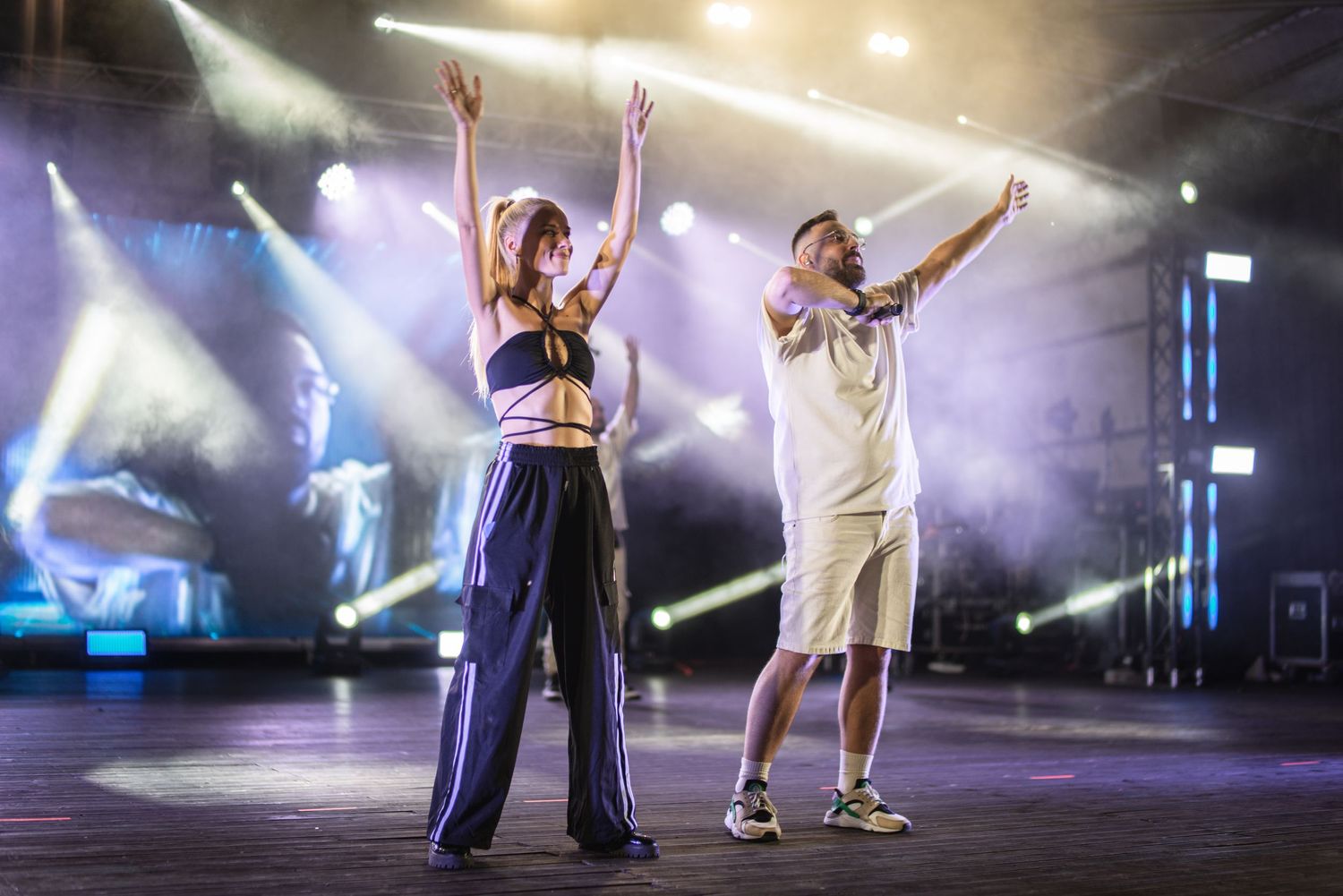 Male and female performers raising arms together on stage under colorful concert lighting.