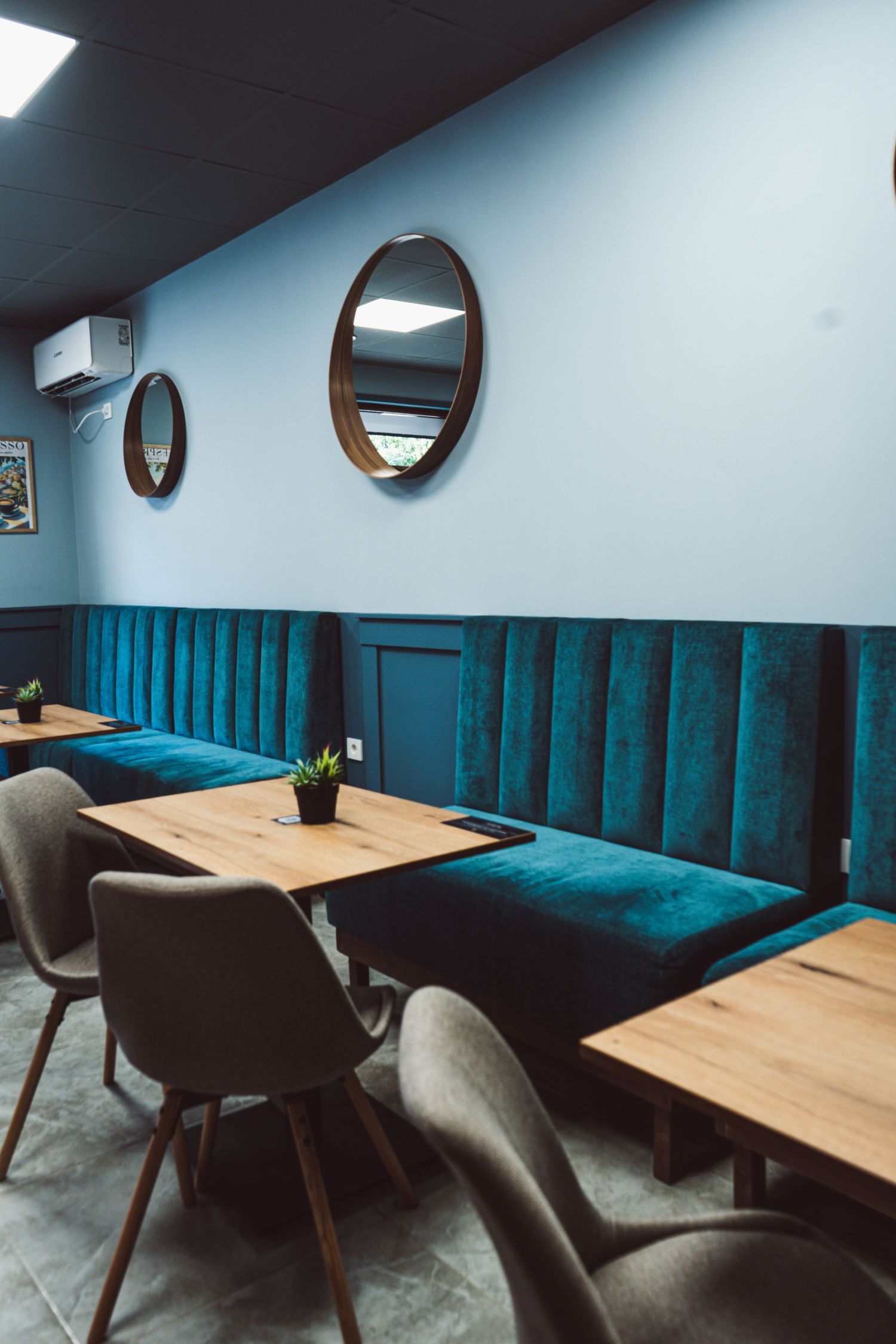 Cozy cafe seating area featuring teal velvet booth, wooden tables, round wall mirrors, and grey chairs.