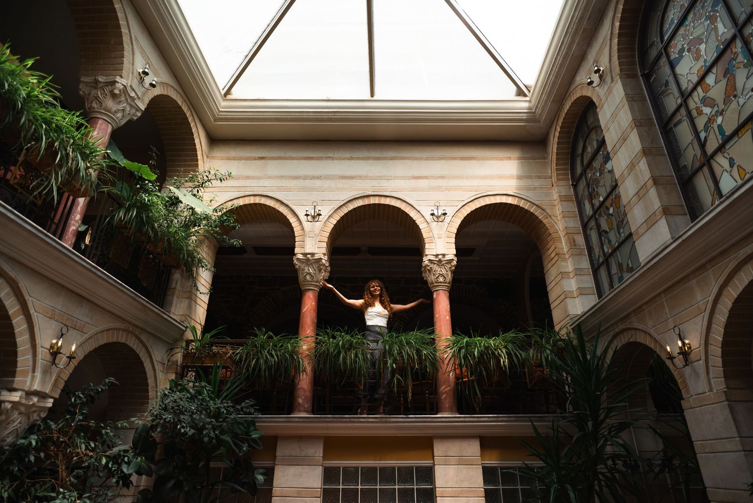 Woman standing on balcony of elegant building with arched columns and lush green plants under skylight.