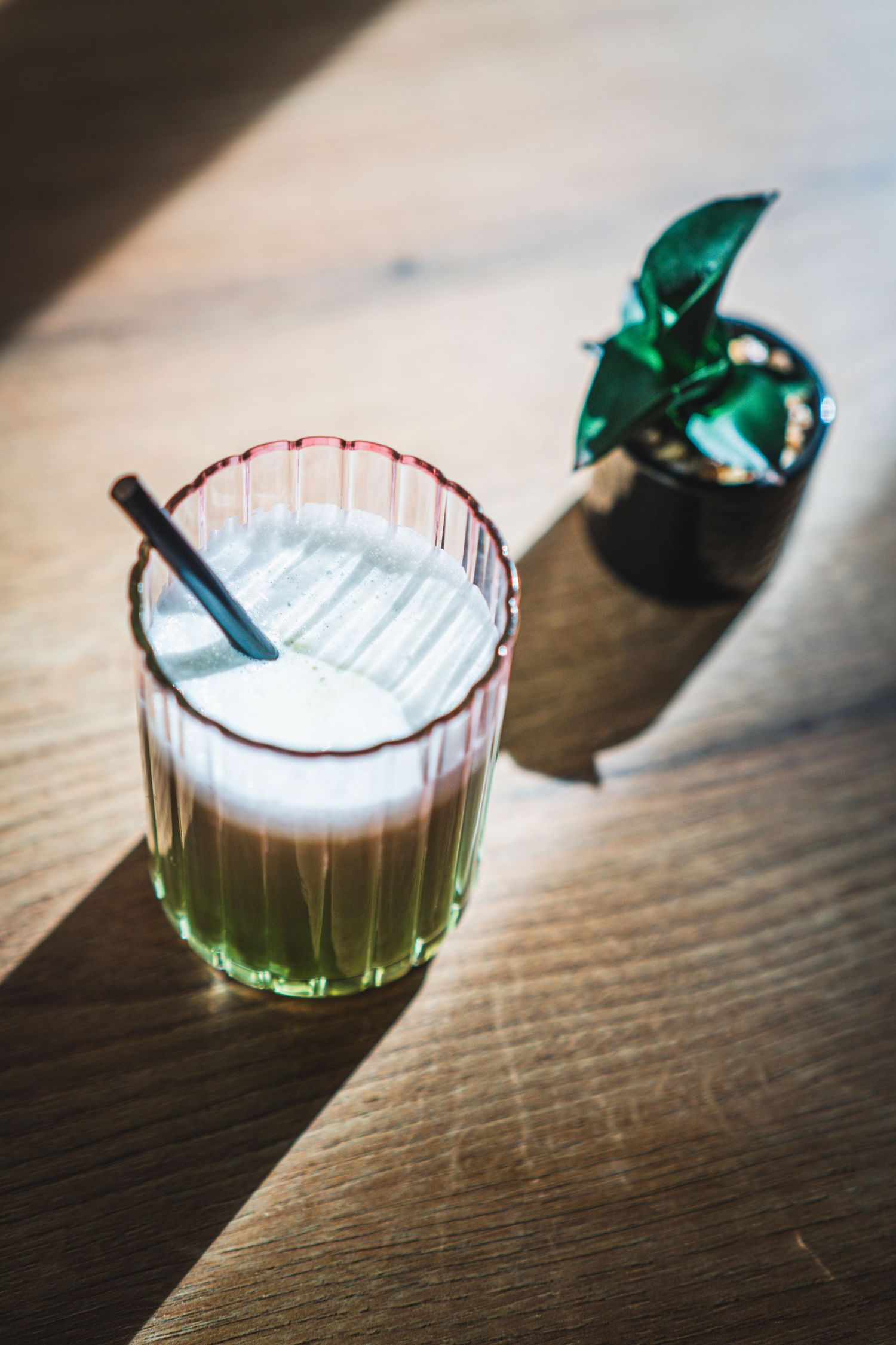 Green matcha latte in ribbed glass with black straw beside small potted plant on wooden surface.
