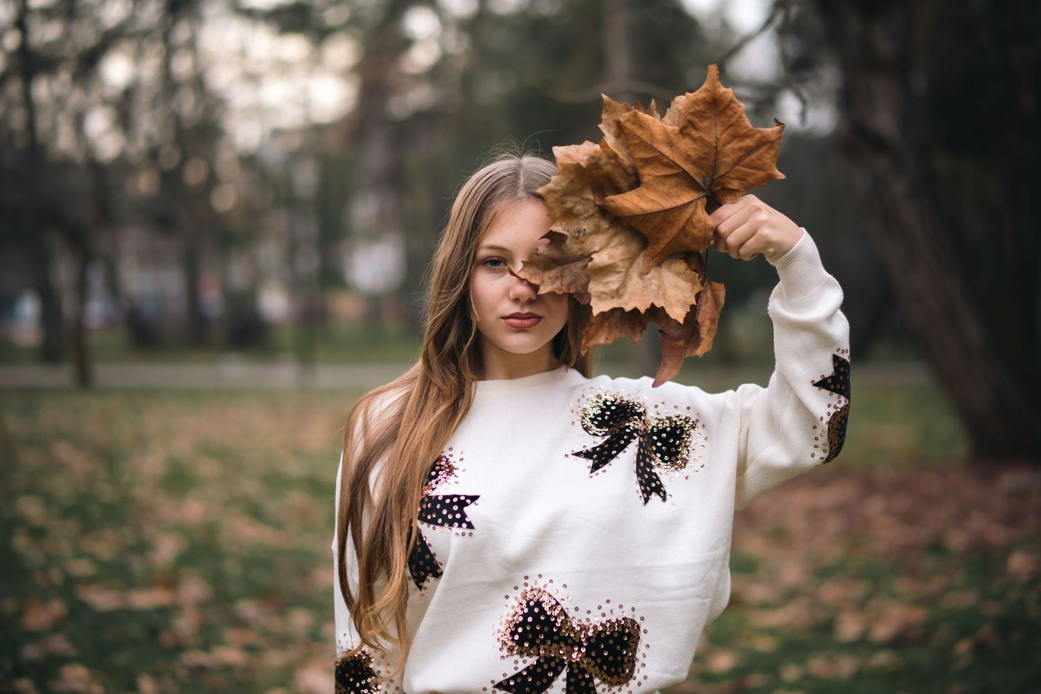 Young woman in white bow sweater holding autumn leaves in a park during fall season.