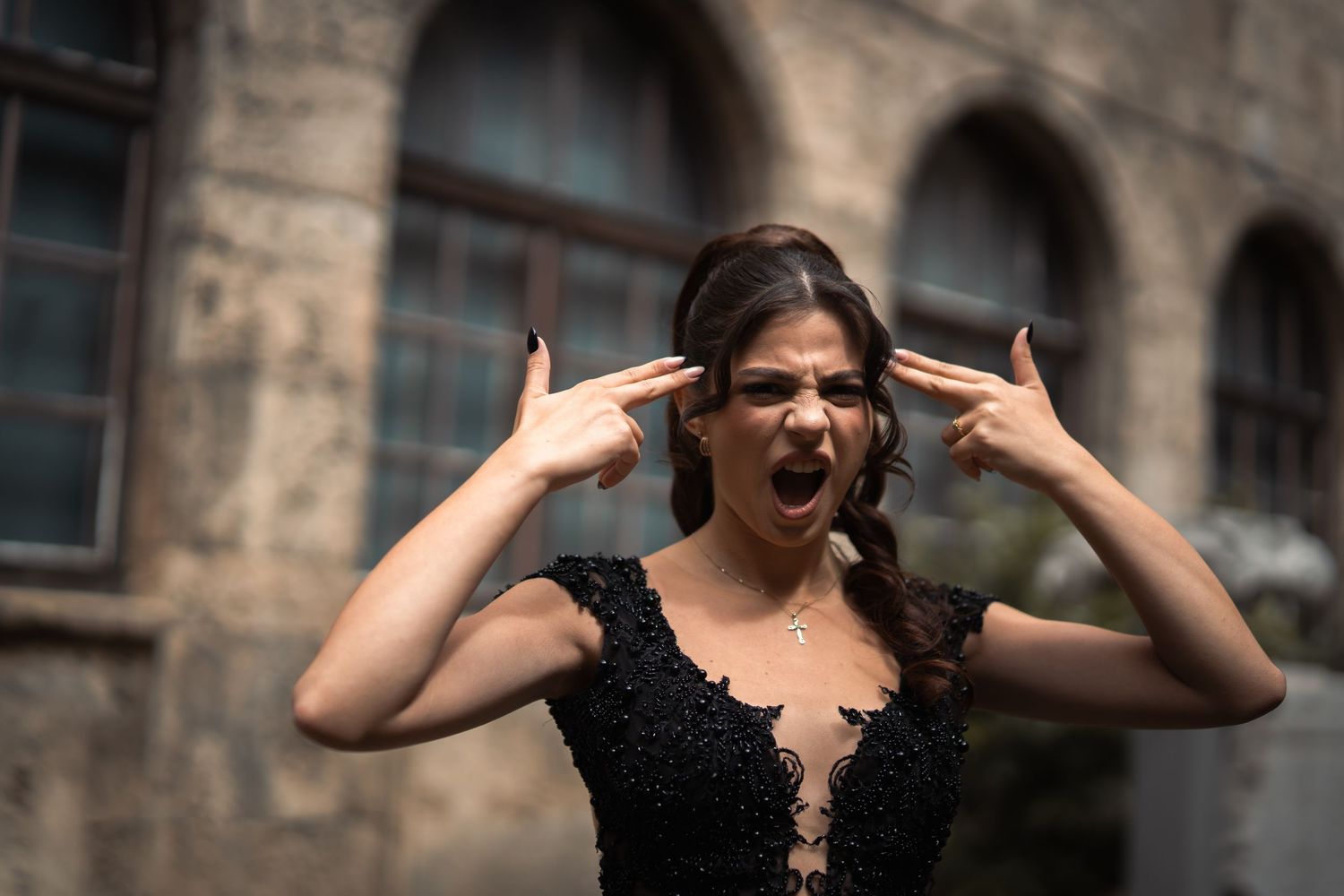 Expressive woman in black lace dress making finger gun gesture to head outdoors.