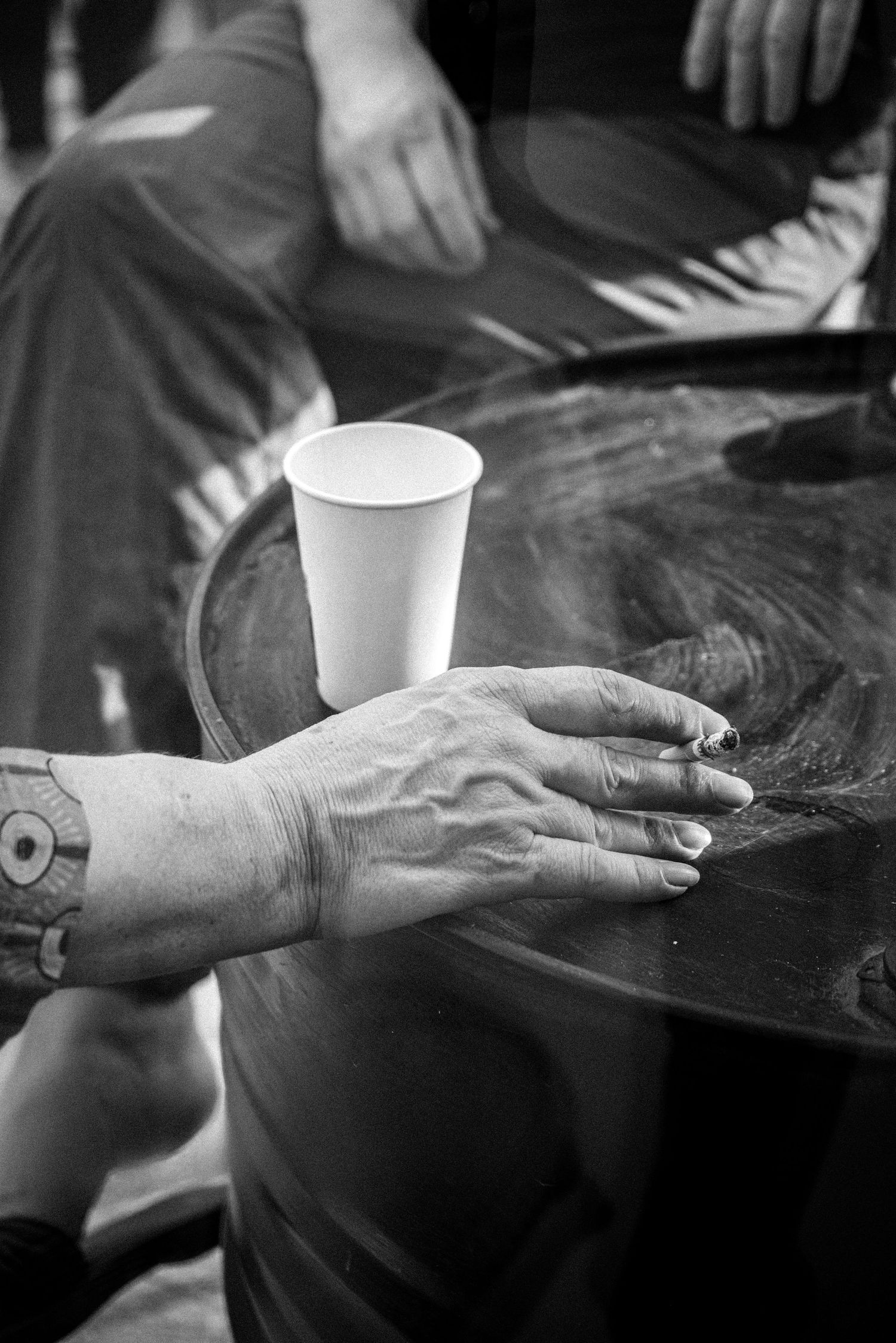 Black and white photo of tattooed hands resting on a table with a white paper cup nearby.