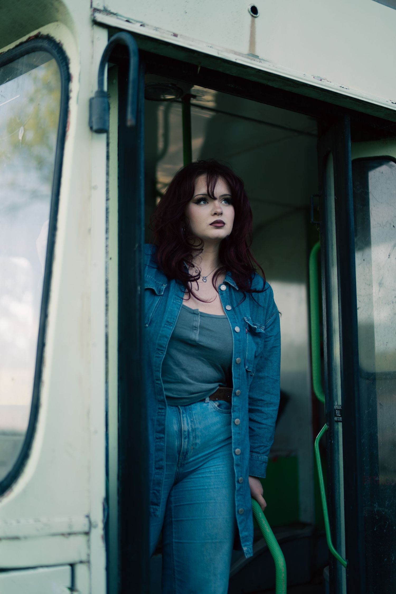 Edgy woman in all-denim outfit stands in doorway of vintage green tram with bold dark lipstick.