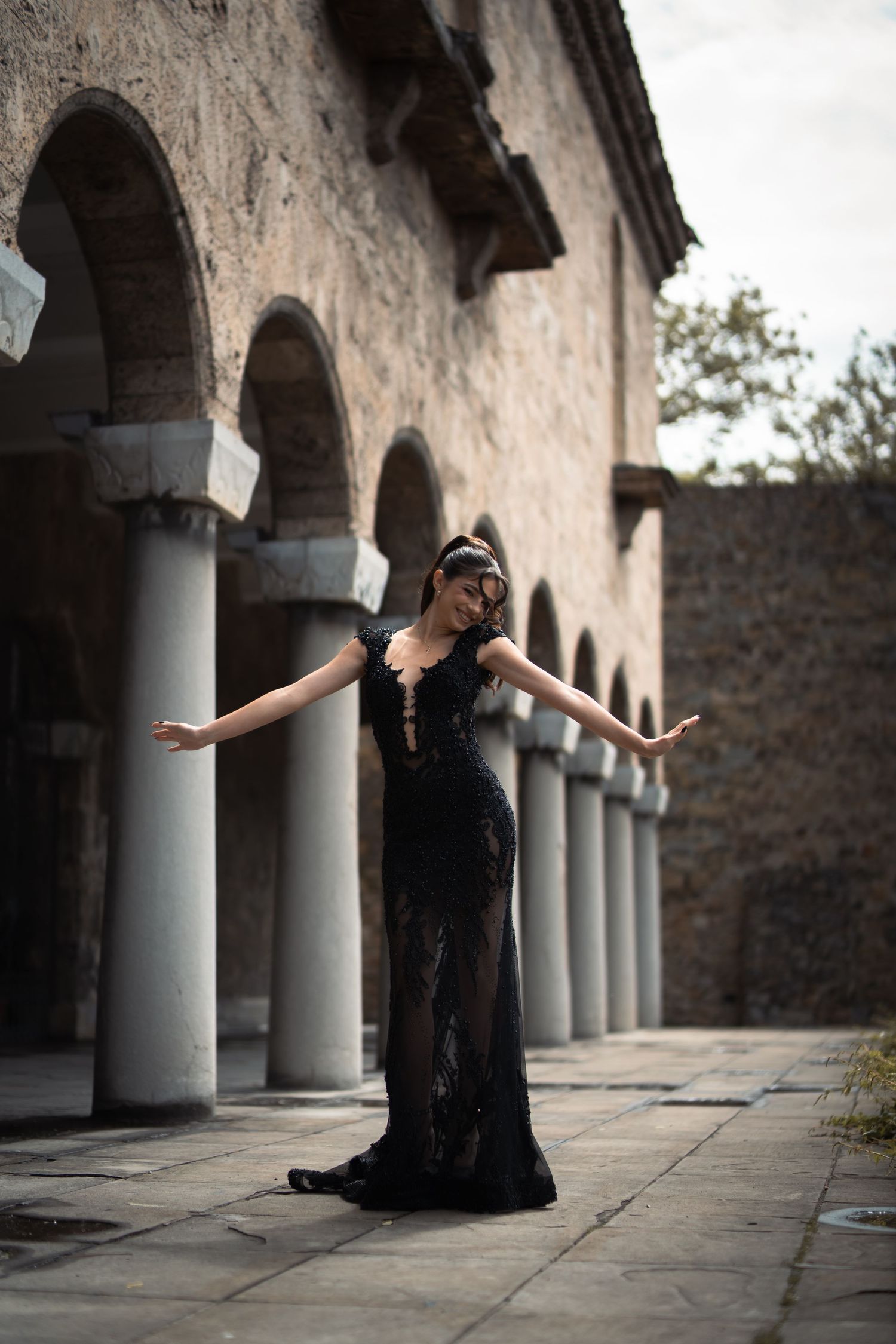 Woman in elegant black sequin gown posing gracefully in front of historic stone arched building.