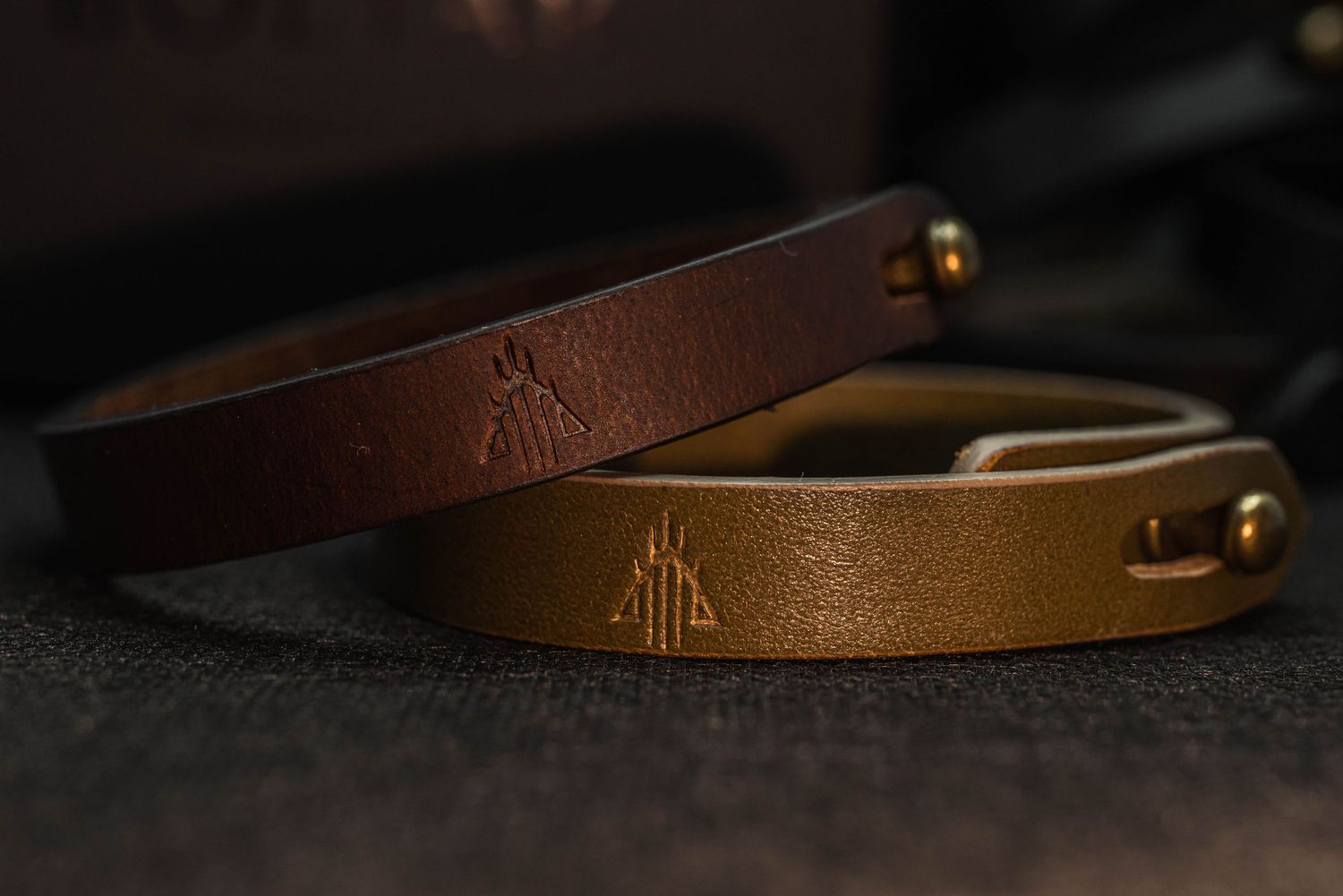 Two handcrafted leather bracelets in brown and gold with embossed logo design on dark textile surface.