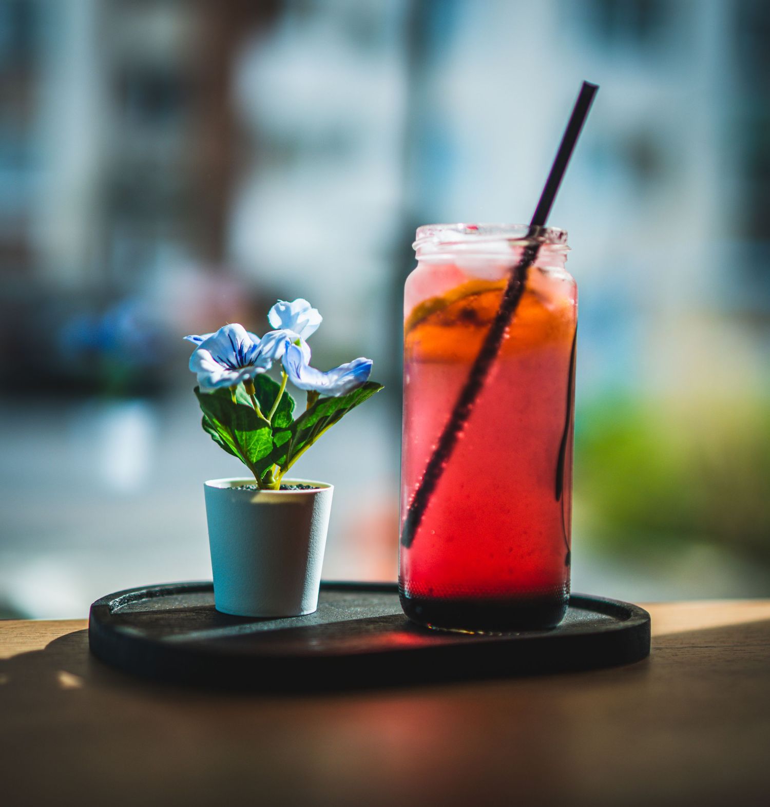 Red iced cocktail drink with black straw beside small blue flower pot on dark tray near window.
