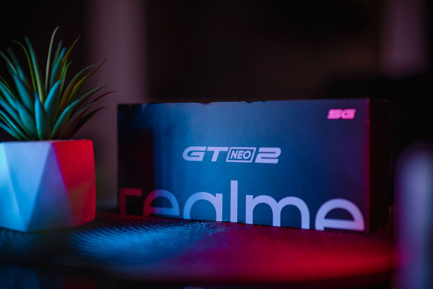 Realme GT Neo 2 5G smartphone box illuminated with colorful neon lights beside a small plant.
