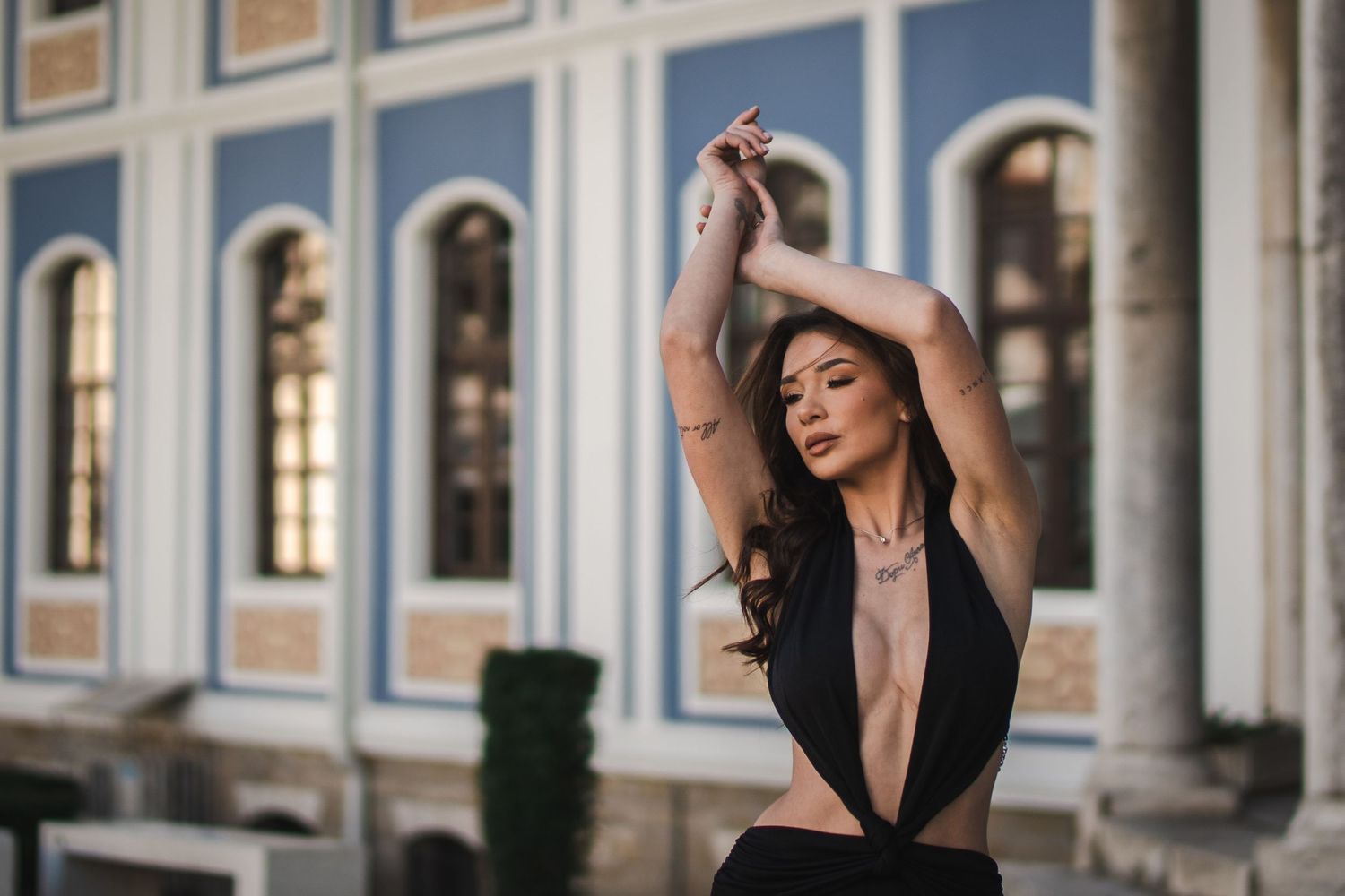 Elegant woman in deep black plunging neckline dress poses with arms raised before ornate blue and white building.