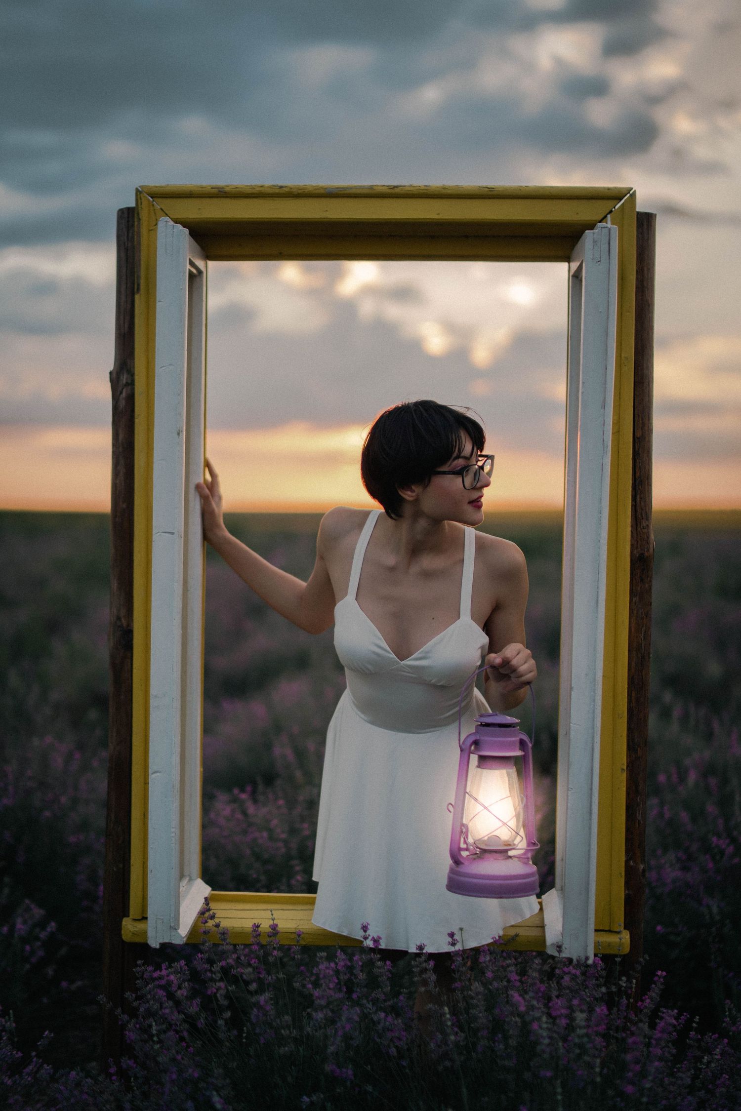 Woman in white dress holds glowing lantern while standing in vintage yellow door frame in lavender field at dusk.
