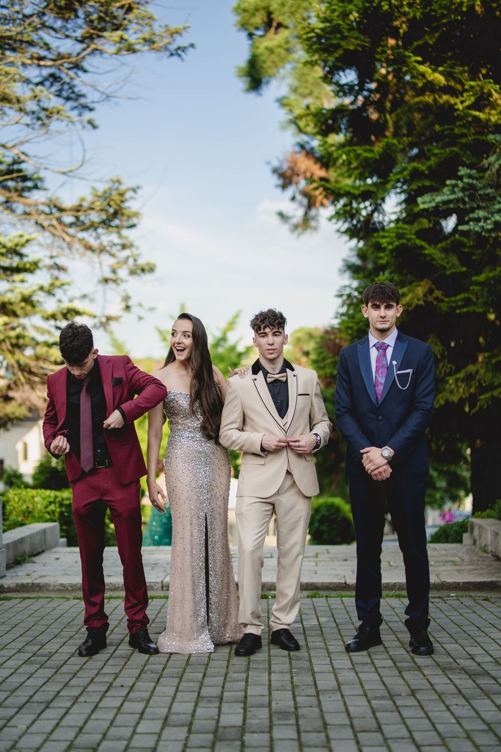 Four teenagers dressed in formal prom attire pose outdoors on a stone path surrounded by lush green trees.