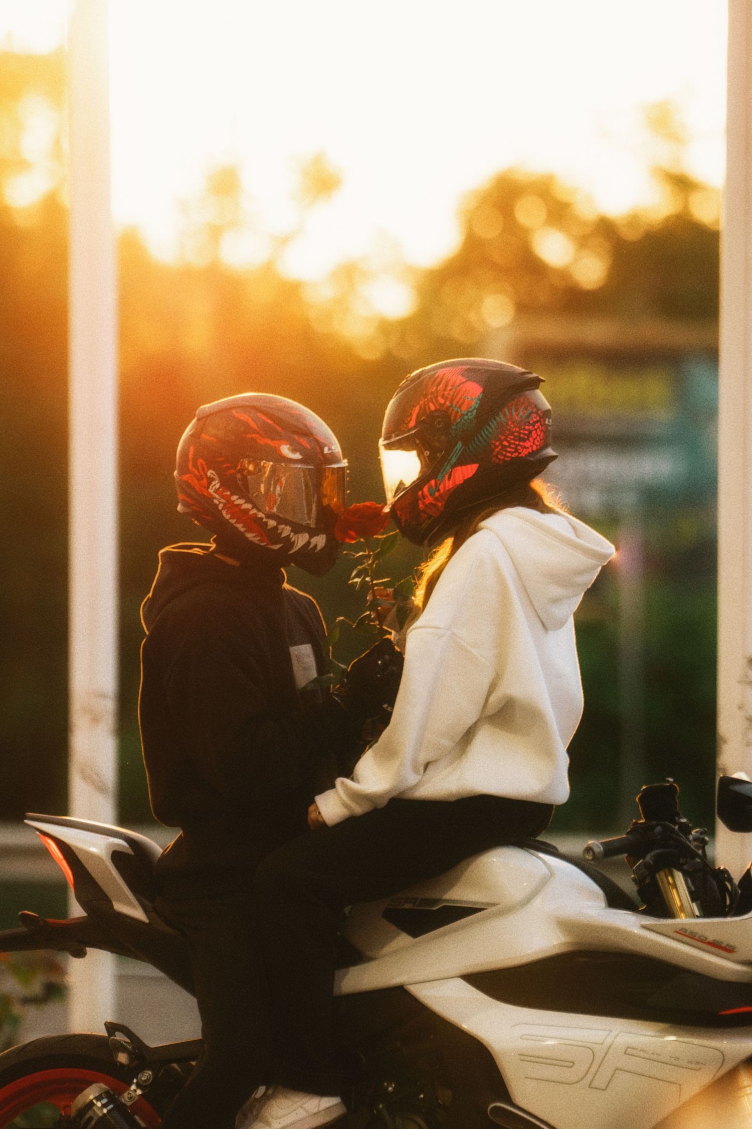 Couple wearing matching red helmets sit intimately on white motorcycle during golden sunset.