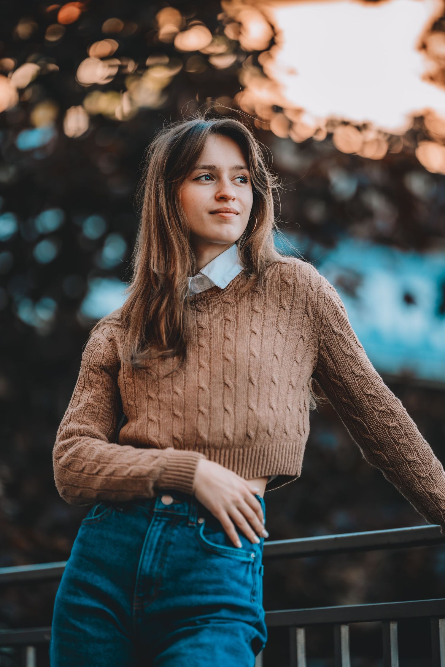 Young woman in beige cable-knit sweater and jeans leaning on a railing at golden hour outdoors.