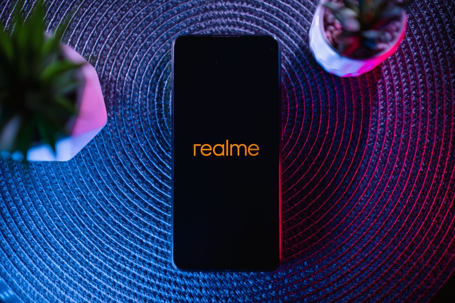 Realme smartphone displayed on a woven surface with colorful neon lighting and small potted plants.