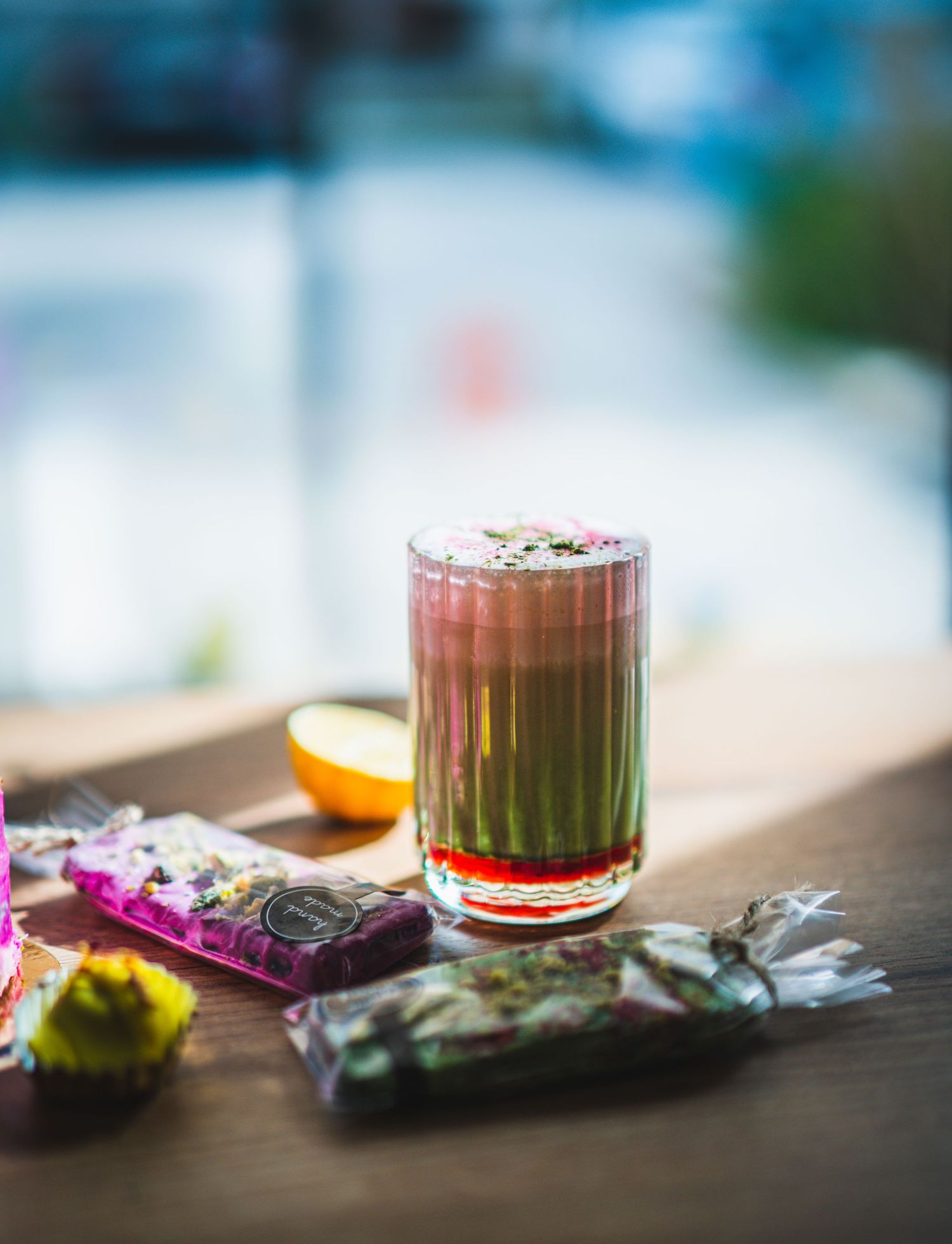 Colorful layered matcha drink in tall glass with candy wrappers and citrus fruit on wooden table.