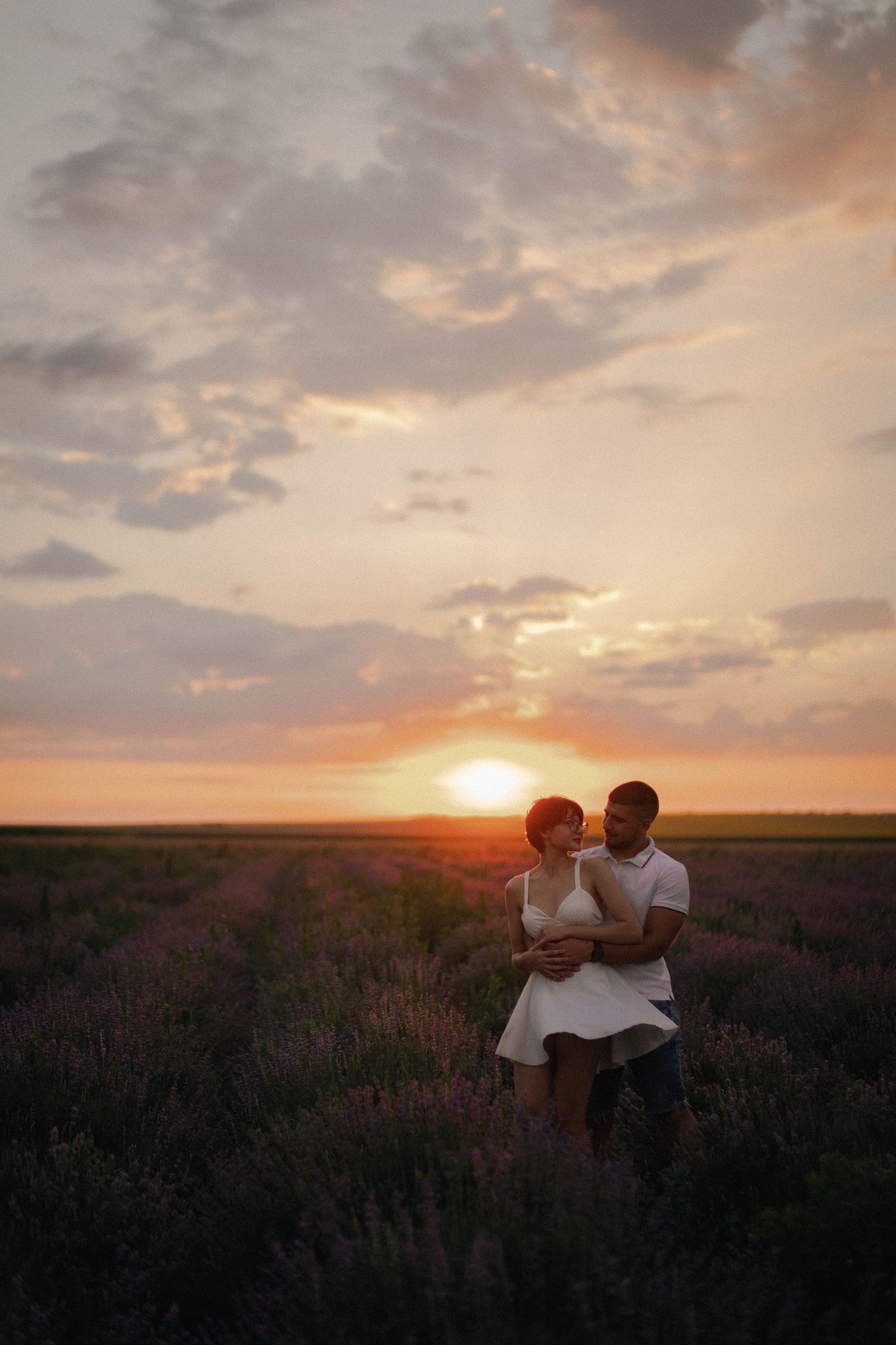 Romantic couple embraces in blooming lavender field under dramatic golden sunset sky.
