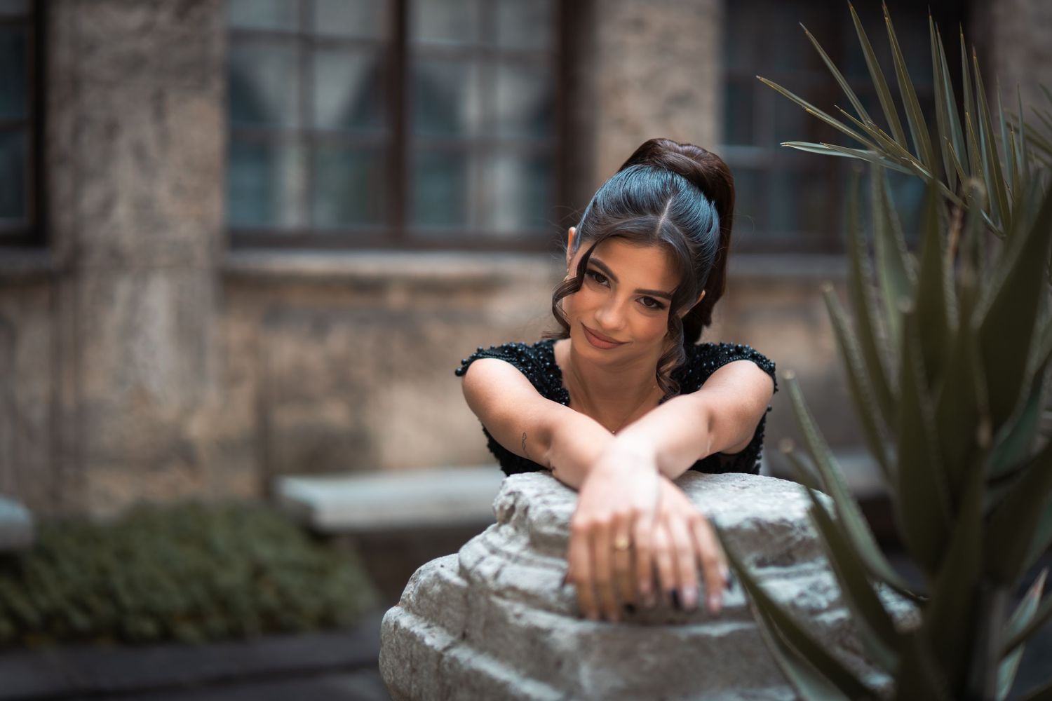 Smiling woman with updo hairstyle leaning on stone sculpture outdoors near tropical plants.