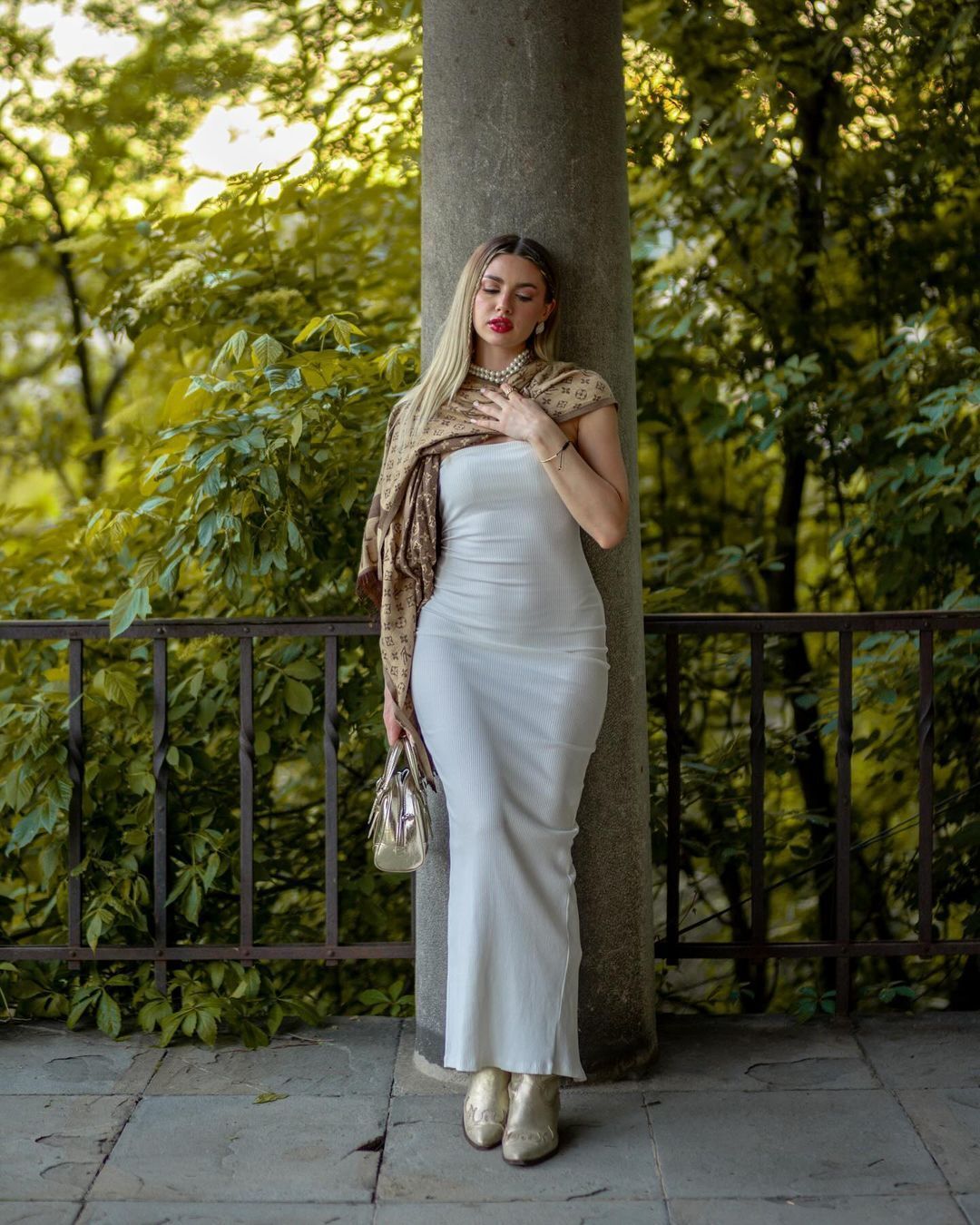 Blonde woman in white slip dress with leopard scarf leaning against stone column in lush park.