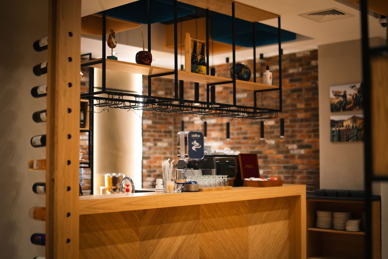 Stylish coffee bar counter with overhead glass rack, brick wall, and espresso machine in cafe interior.