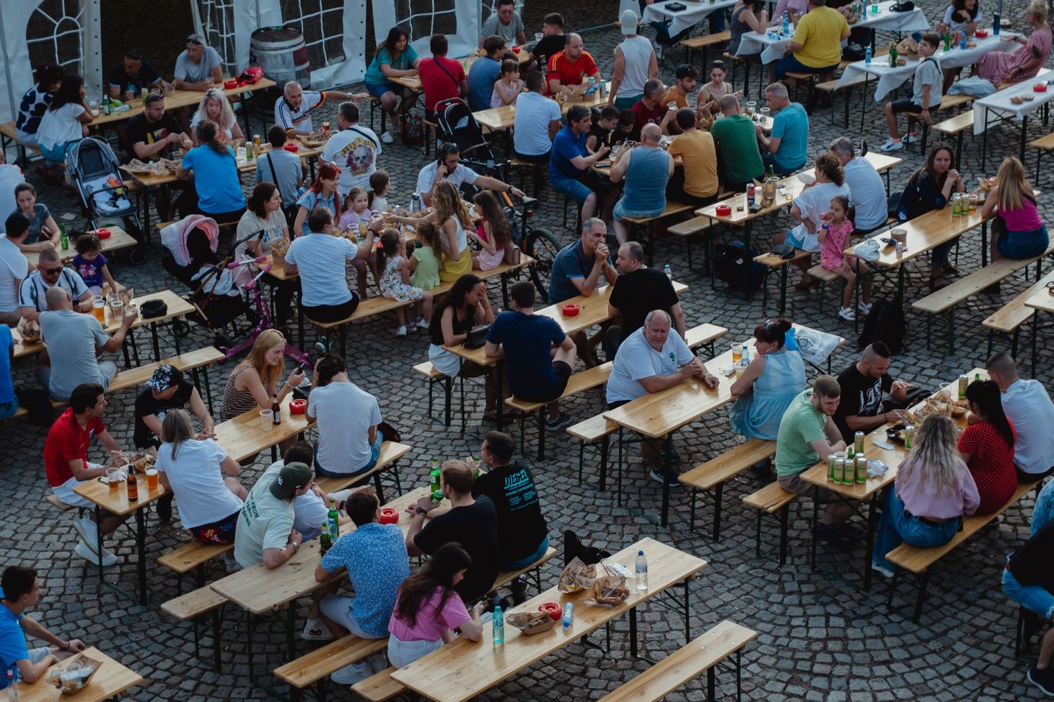 Aerial view of crowded outdoor beer garden with people sitting at wooden benches on cobblestone plaza.