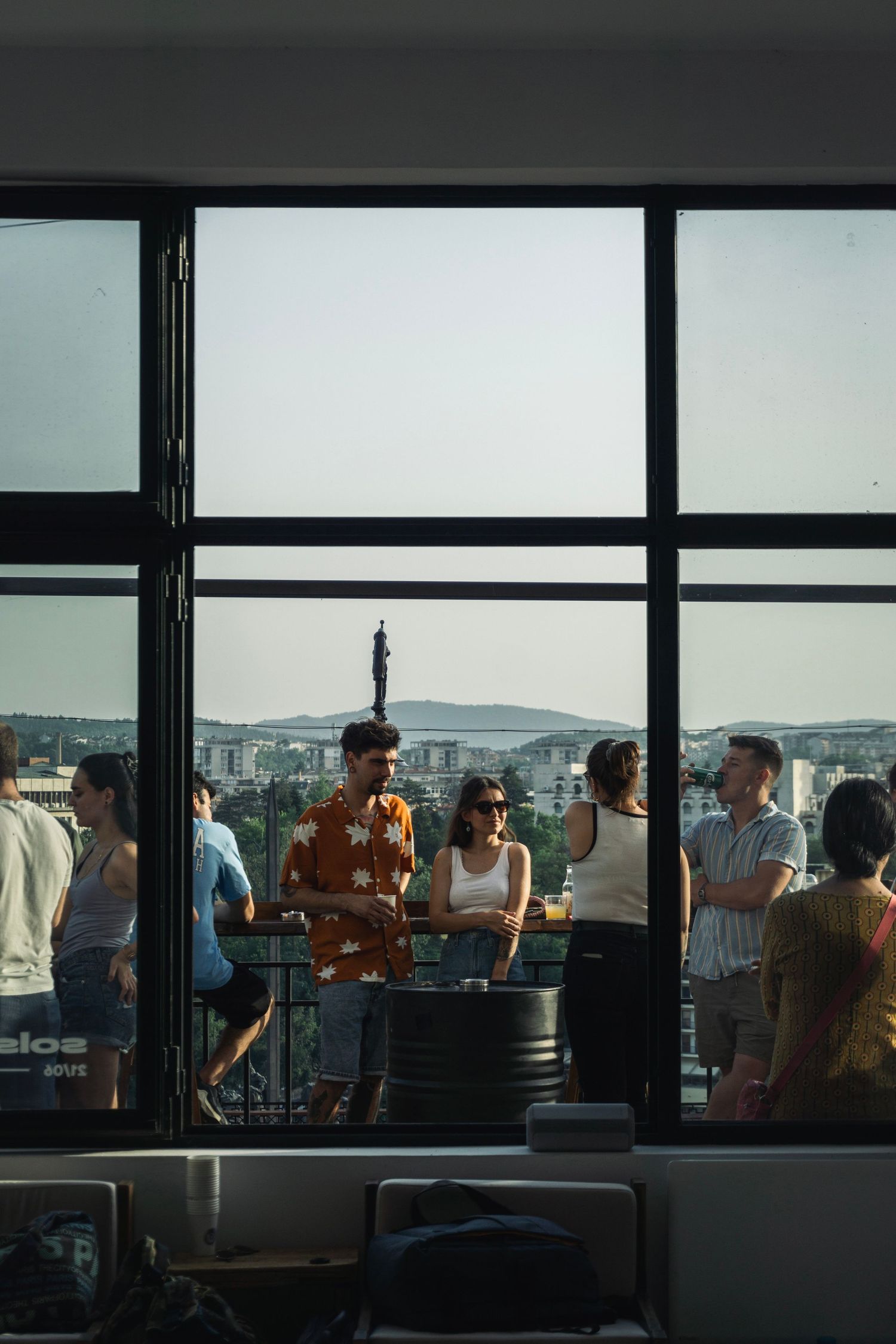 People mingling on a rooftop terrace with a panoramic city skyline view through large glass windows.