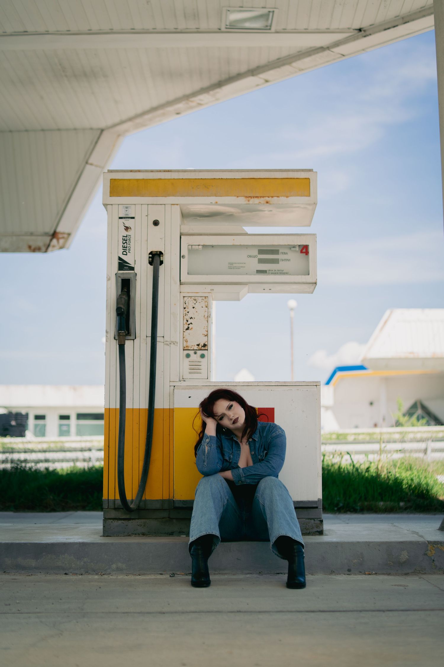 Woman in denim outfit sitting against yellow gas pump at abandoned gas station.