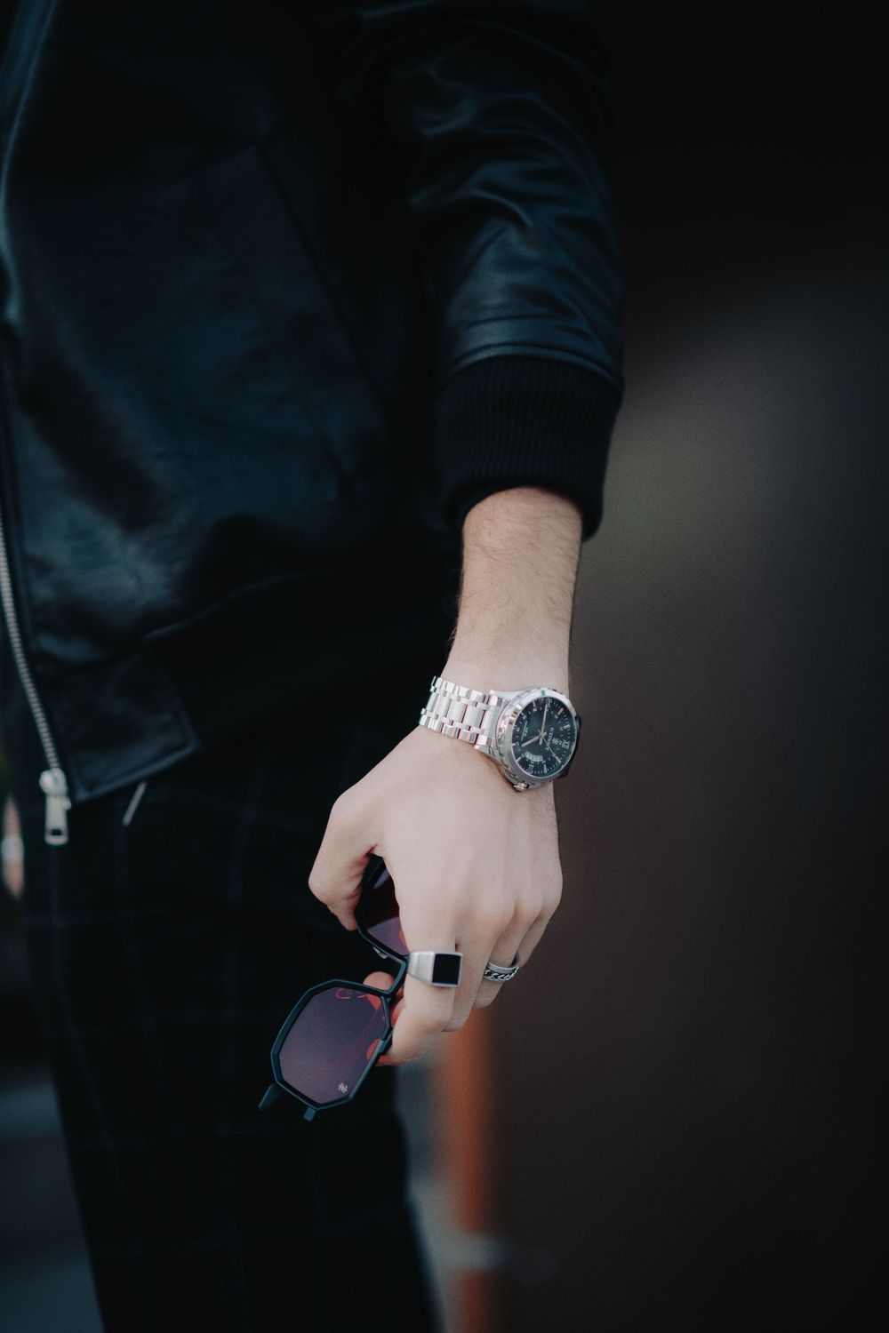 Man in black leather jacket holding sunglasses with a silver watch and ring on his hand.