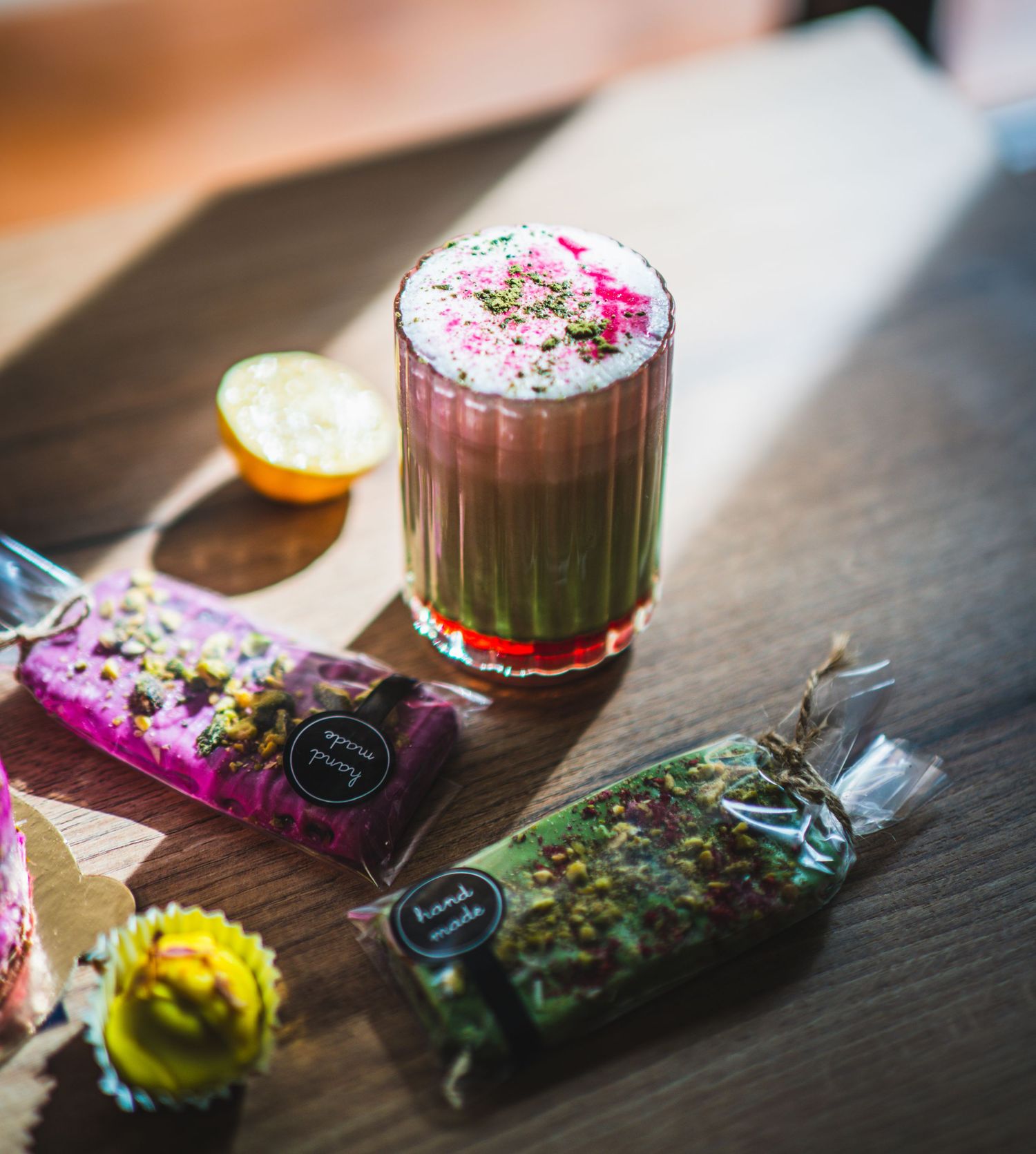 Artistic matcha drink with pink floral foam topping beside herb-filled candy packages on wooden table.