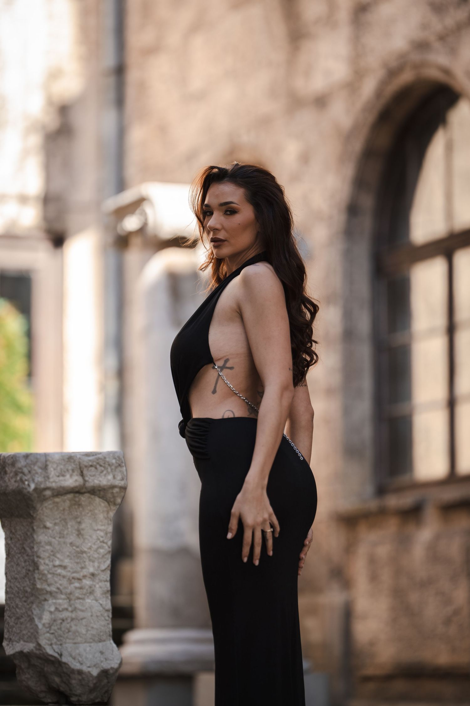 Tattooed woman in black backless dress posing beside stone column of historic building.