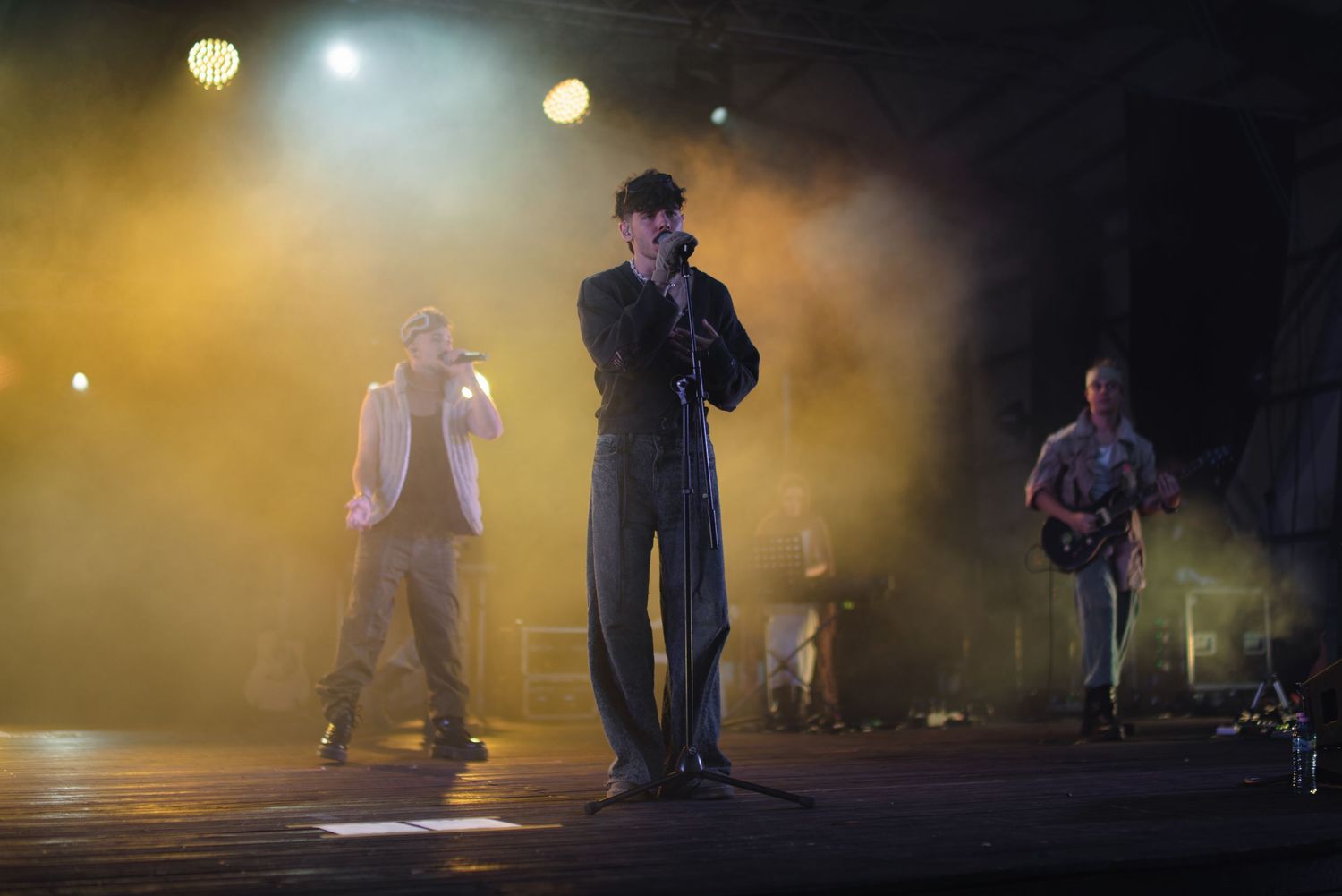 Three musicians performing on stage under dramatic yellow fog lights at outdoor concert.