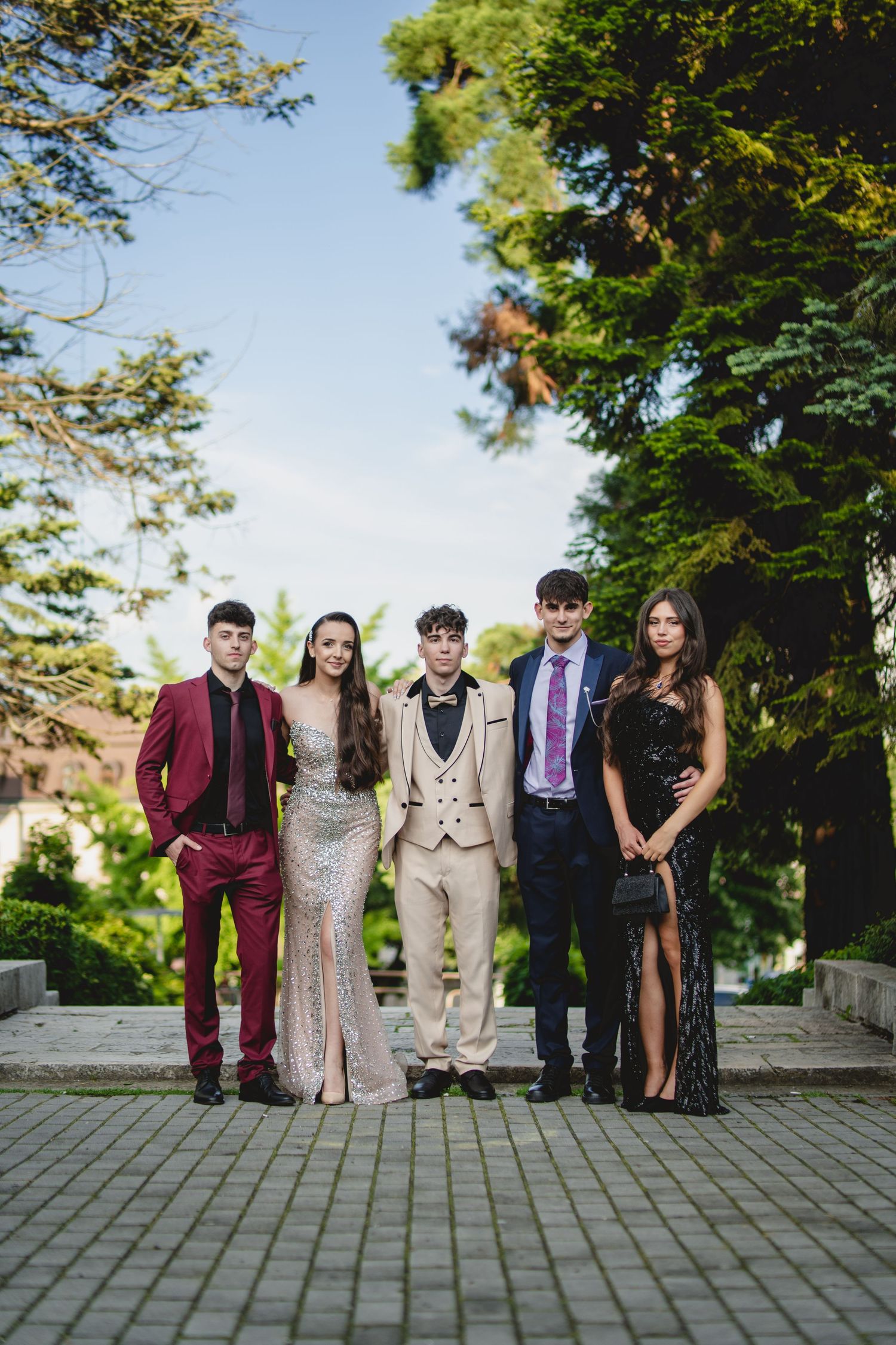 Group of five teens in formal prom attire posing outdoors among tall green trees.