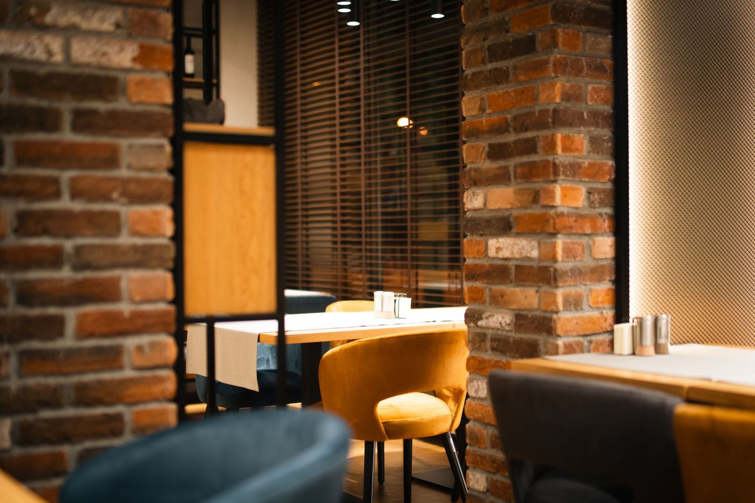 Cozy cafe interior with exposed brick walls, yellow chair, wooden blinds, and warm ambient lighting.