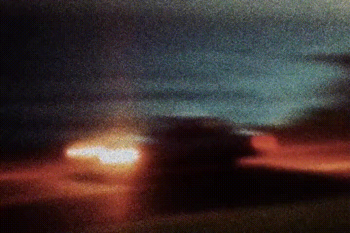 Blurry nighttime photo of a car with glowing headlights on a dark road under a dusky sky.