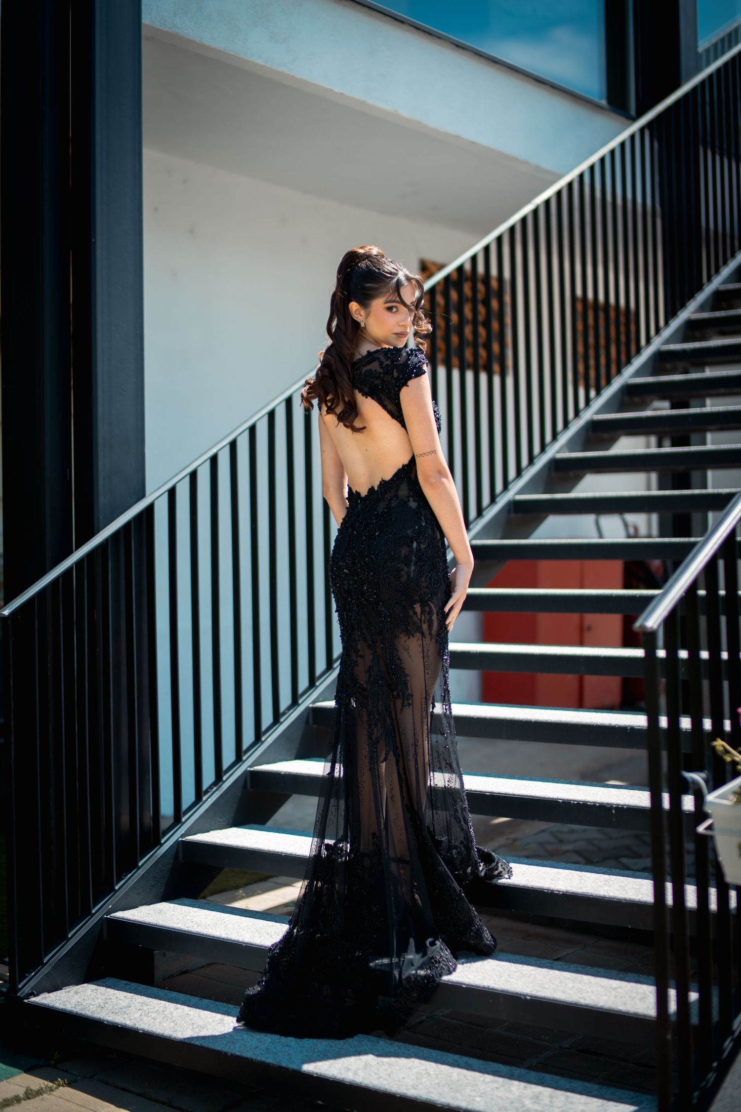 Woman in elegant backless black lace mermaid gown posing on modern staircase in sunlight.