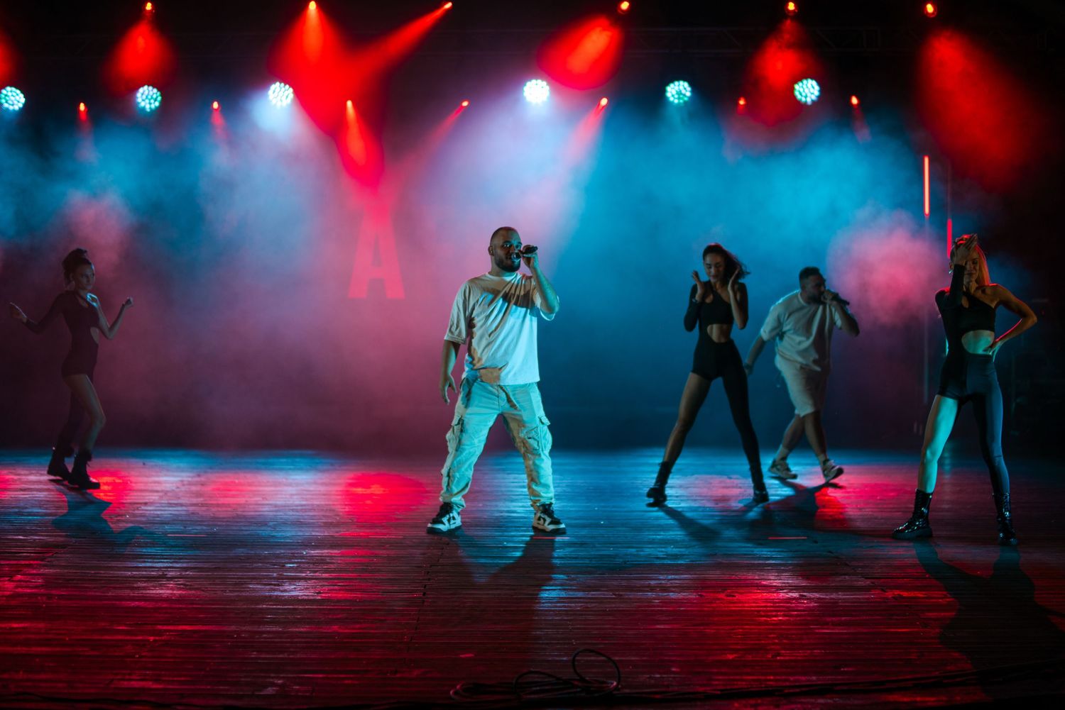 Hip-hop artist performing with backup dancers under red and blue stage lights with smoke effects.