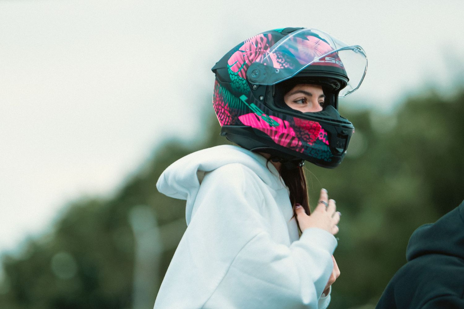 Person wearing colorful pink and teal motorcycle helmet outdoors with trees in background.