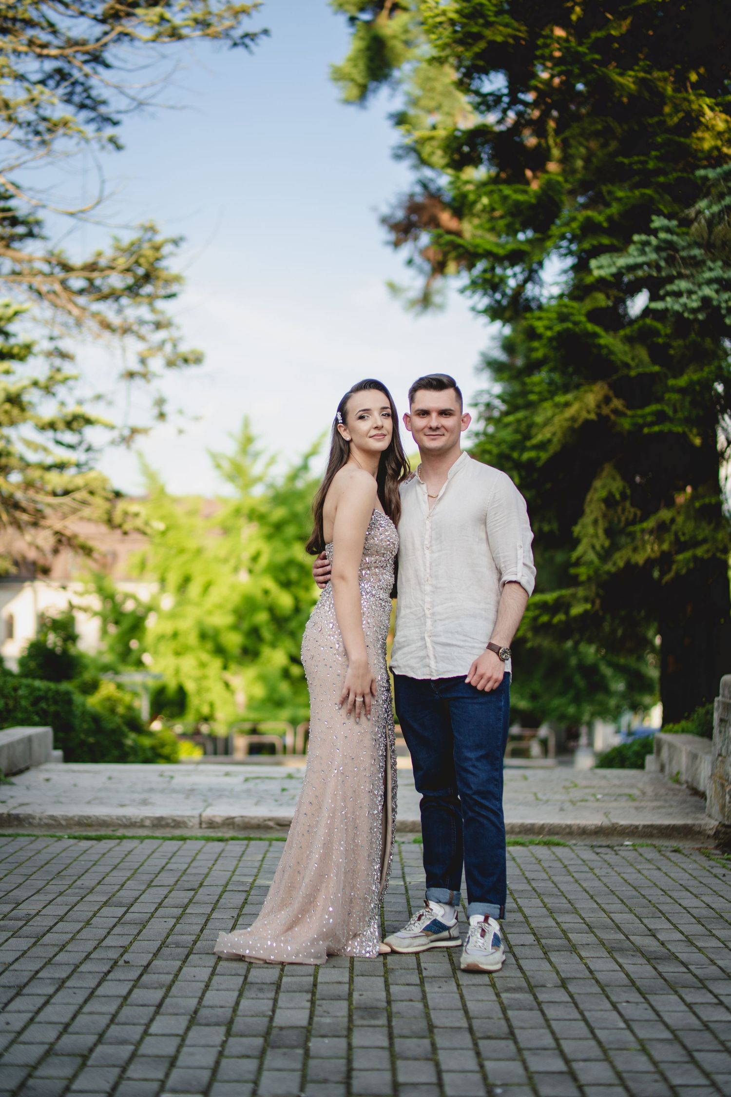 Young couple posing outdoors, woman in elegant sequin gown, man in casual white shirt and jeans.