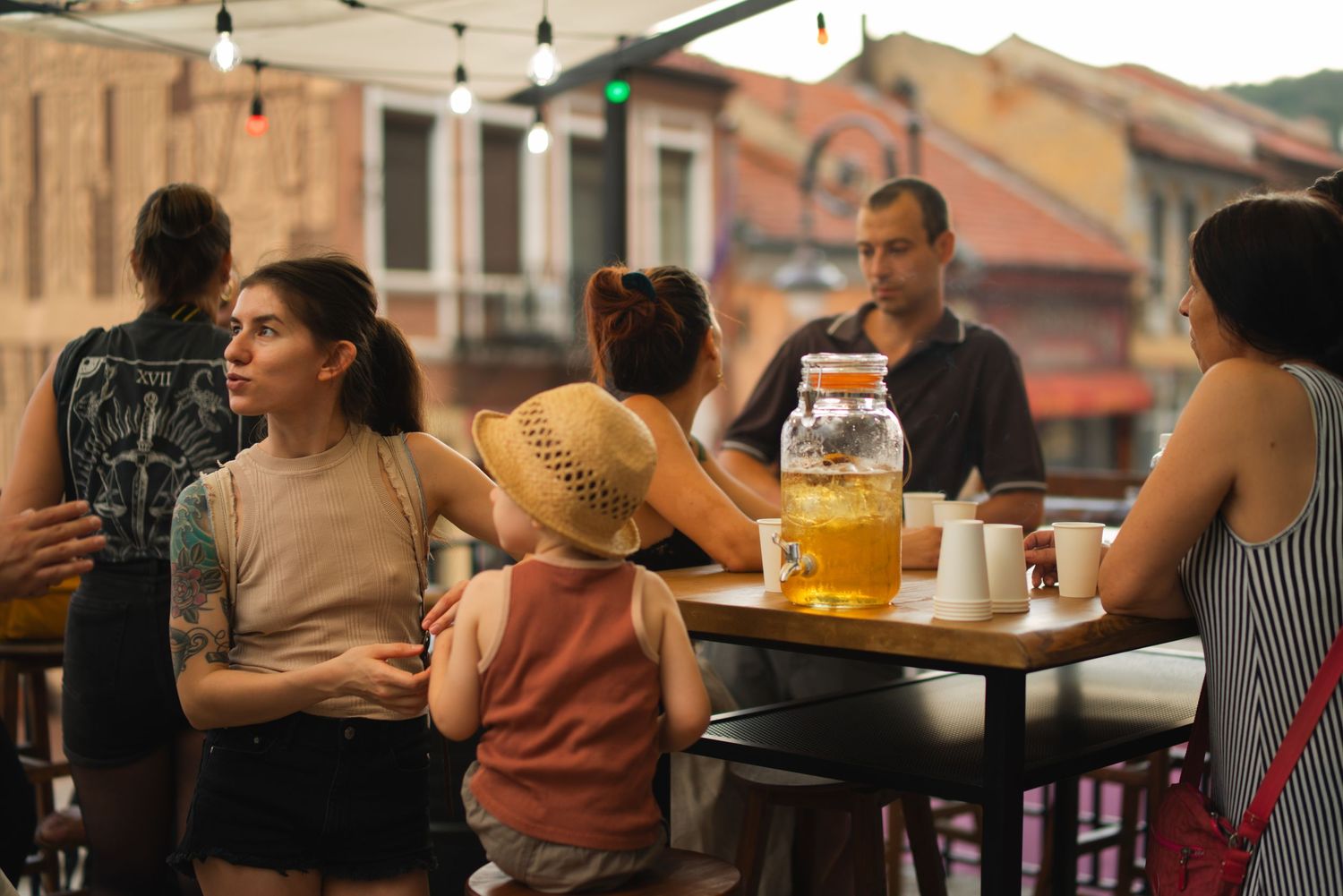 Group of people relaxing at outdoor cafe terrace with large jar of iced tea on table at dusk.
