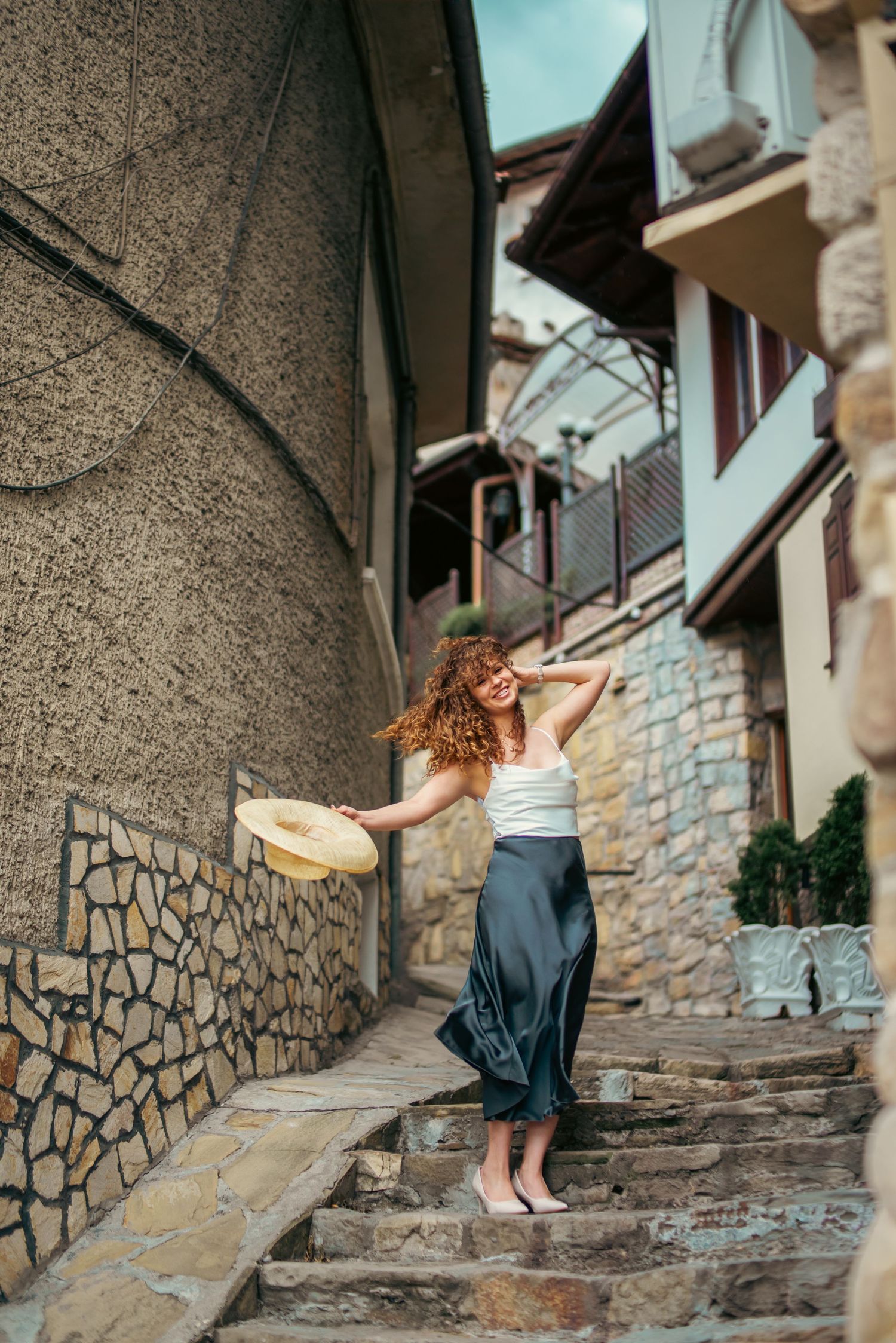 Joyful curly-haired woman in white top and satin skirt twirls hat on stone staircase in old town alley.