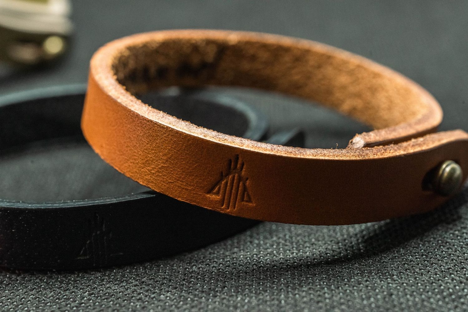 Tan leather bracelet with embossed mountain logo and brass snap closure on dark background.