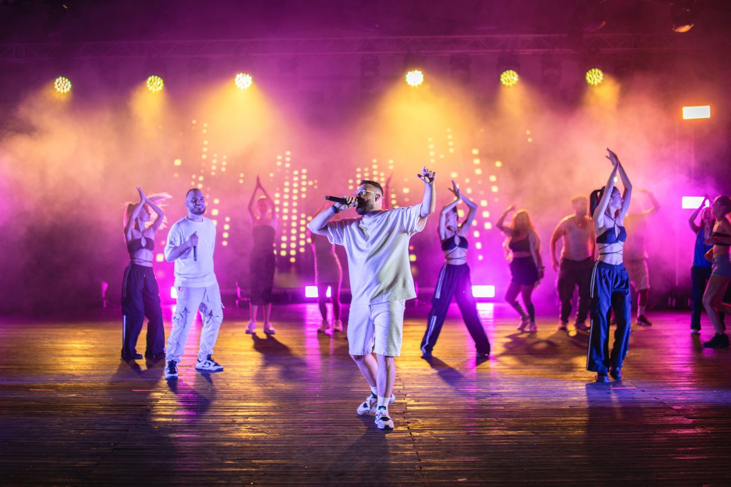 Lead performer with backup dancers on stage surrounded by pink and purple smoky stage lights.