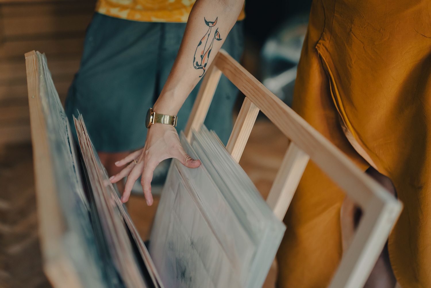 Close-up of tattooed hand browsing through canvas paintings in wooden frames at art studio.
