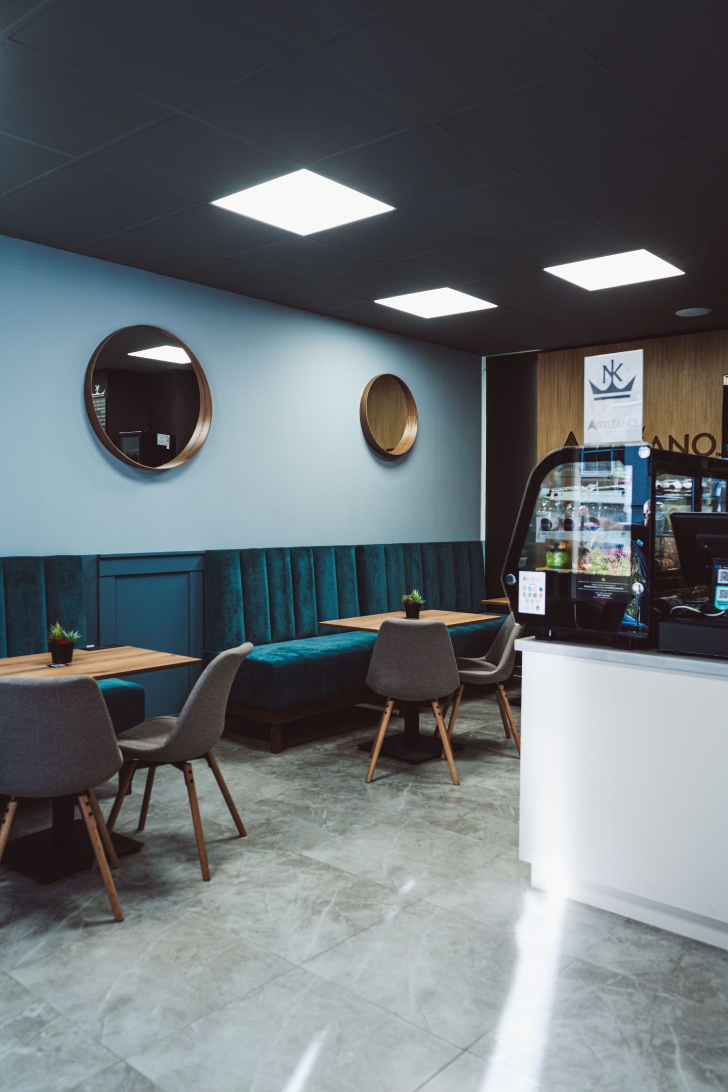 Modern cafe interior with teal upholstered banquette, round mirrors, grey chairs, and white service counter.